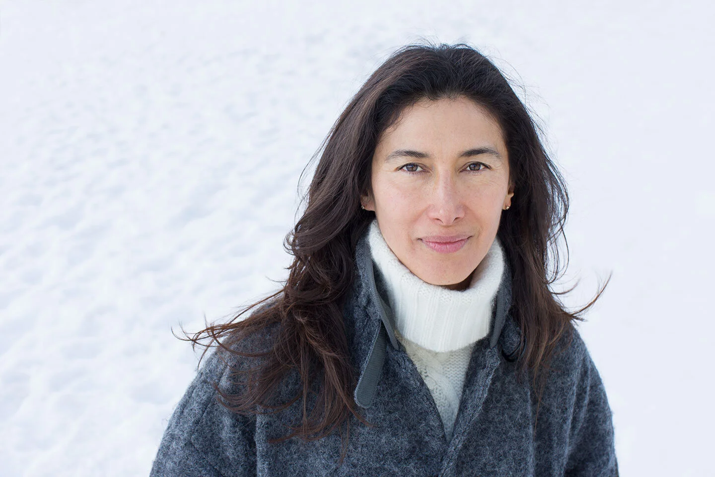 A Mexican woman with dark brown hair and a neutral expression standing outdoors in a snowy environment, wearing a gray coat and a white turtleneck in Lake Tahoe, CA.