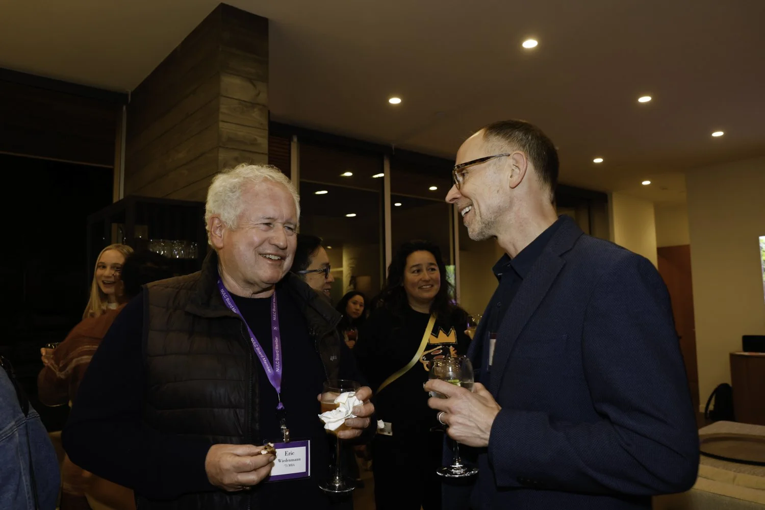 Two men smiling and talking at a social event, holding glasses of wine. One man has white hair, wearing a black vest and a name tag, the other man has short hair, glasses, and a dark blazer. Several women are in the background, one with a yellow sash