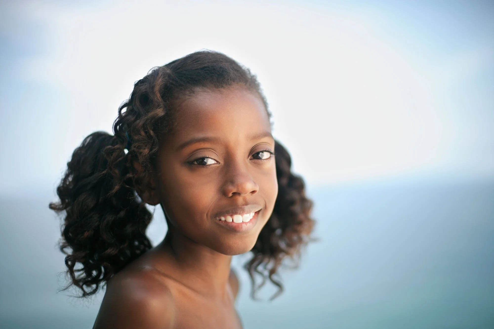 A young Black Brazilian girl with brown curly hair and a slight smile, outdoors with a blurred blue and white background at Ipanema beach in Rio de Janeiro, Brazil.