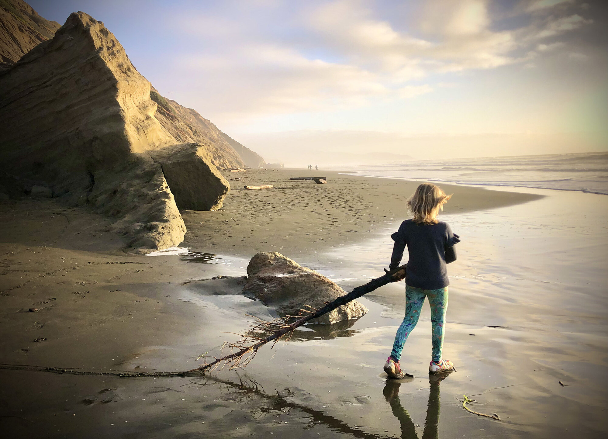 Young girl carrying a long branch in the sand on the beach walking towards the sun at sunset at Fort Funston, San Francisco, CA.