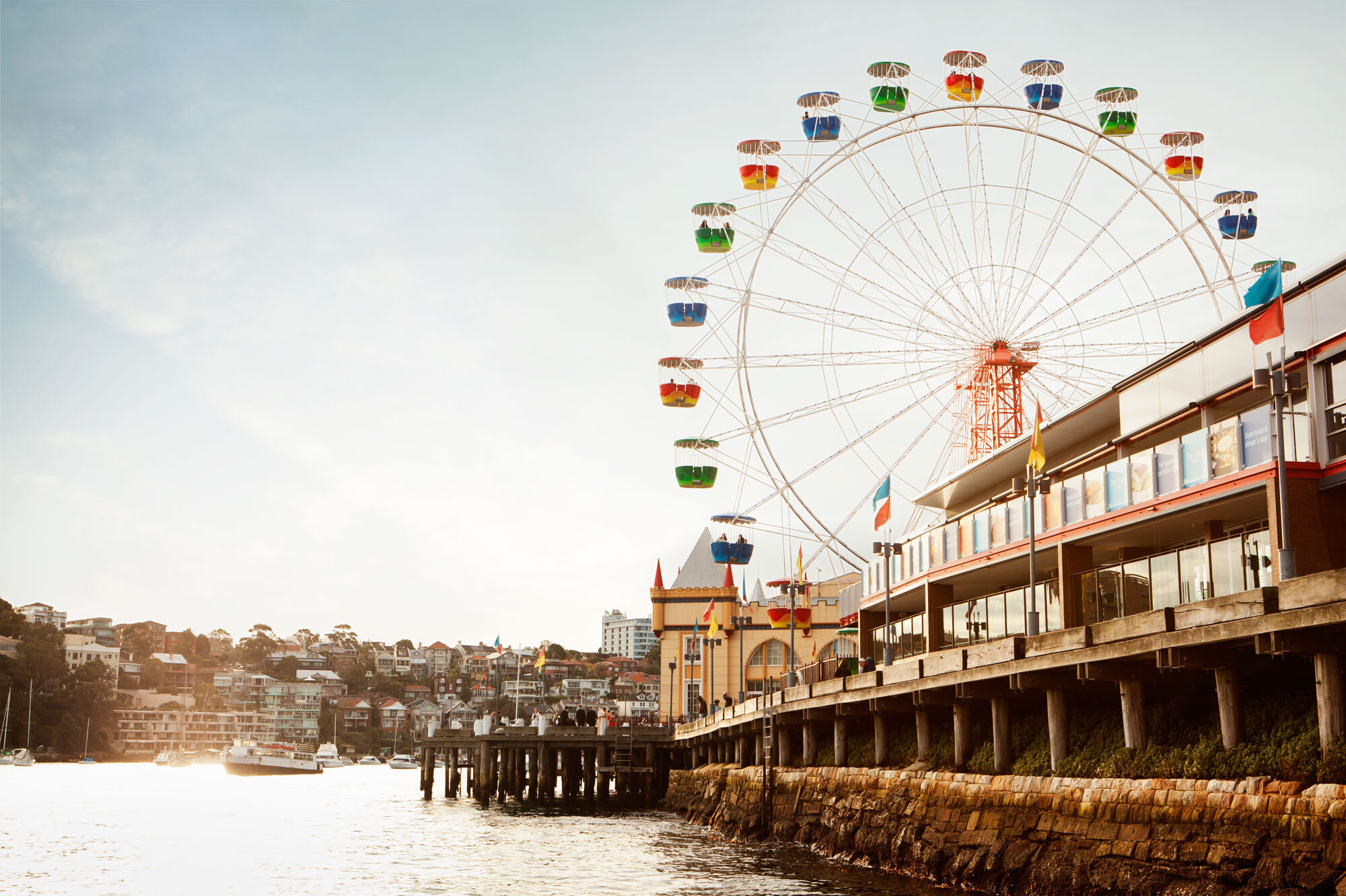 A sunset view of a waterfront pier with a colorful Ferris wheel in the background and a castle-like building. There are boats in the water and people walking on the pier, with flags flying along the railing in Australia.