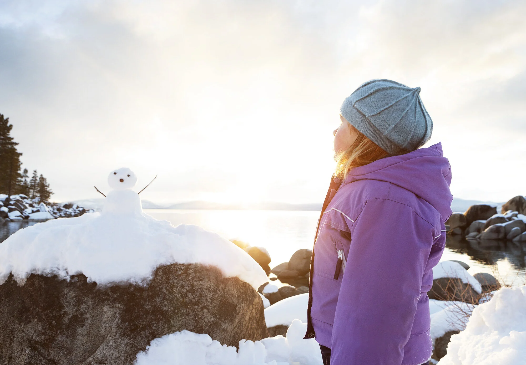 A girl in a purple winter jacket and a gray knit hat looks at a small snowman on a rock, with a snowy landscape and a body of water during sunset in the background at Sand Harbor, Lake Tahoe, CA.