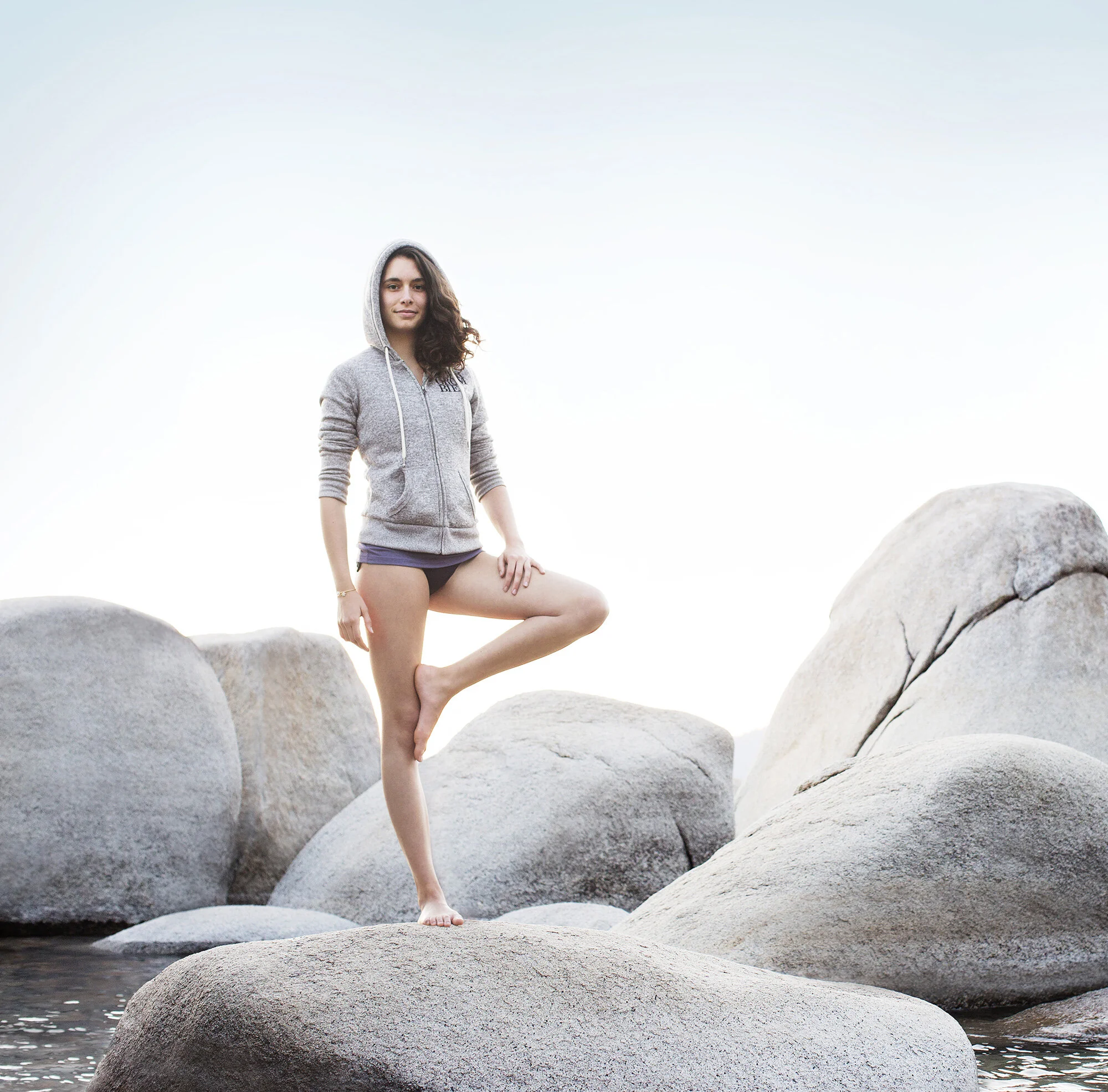 Young woman with curly brown hair standing on rocks by water, wearing a gray hoodie and black shorts, with one leg resting on a rock at Sand Harbor, Lake Tahoe, CA.