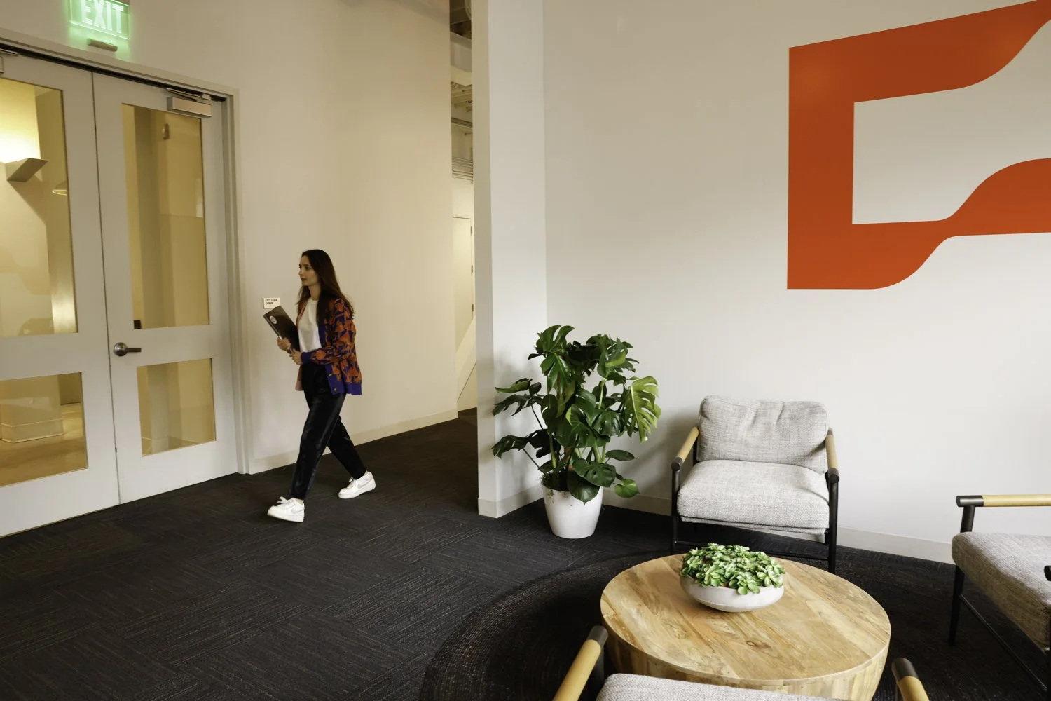 A woman walking in a modern office lobby holding a folder with a laptop sticker, with a white wall, a green potted plant, and a sitting area with chairs and a coffee table in the background Brex headquarter in San Francisco, CA.