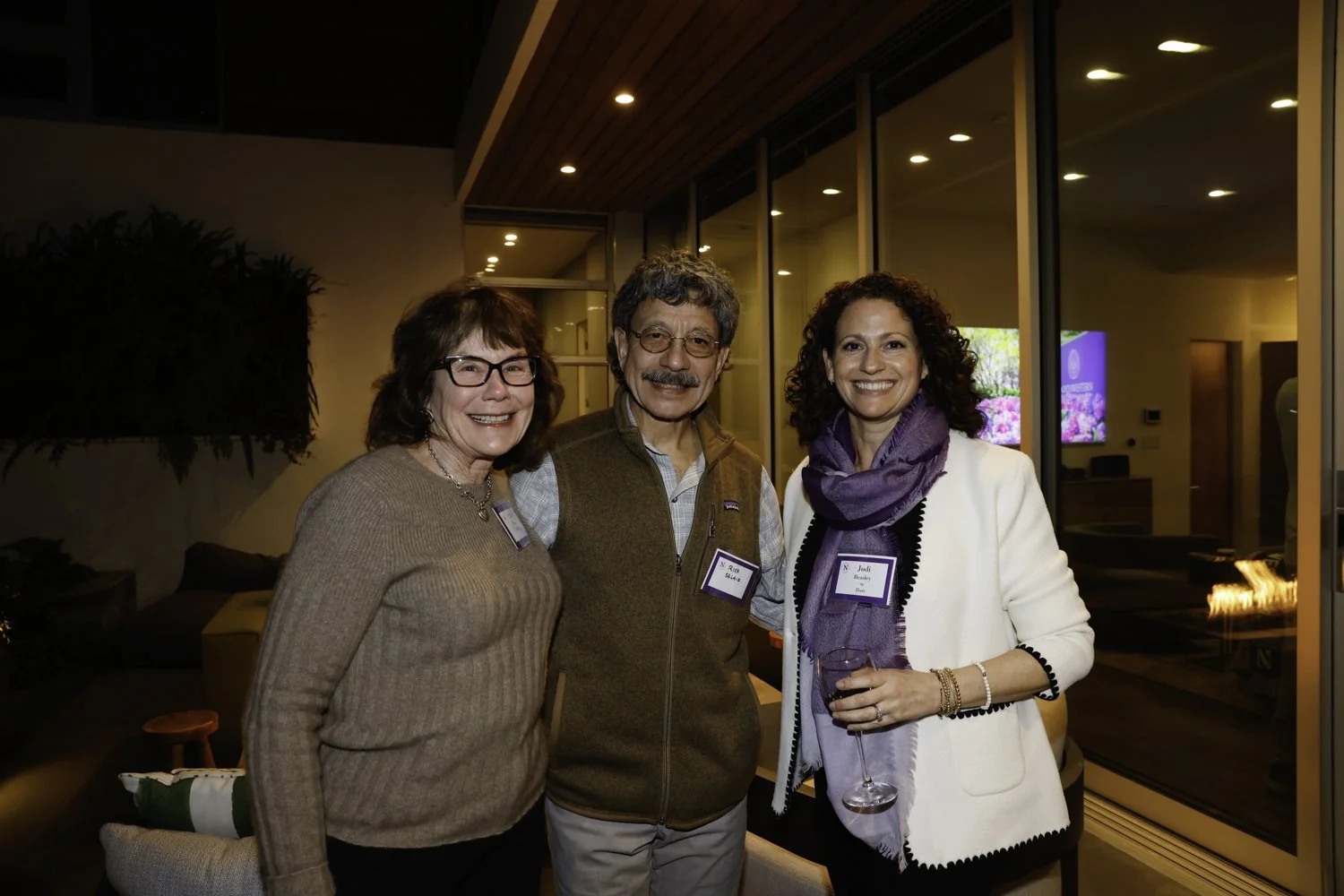 Three people smiling at a social gathering in a modern indoor setting with large windows and a fire feature, one woman holding a glass of wine Brex headquarter in San Francisco, CA.