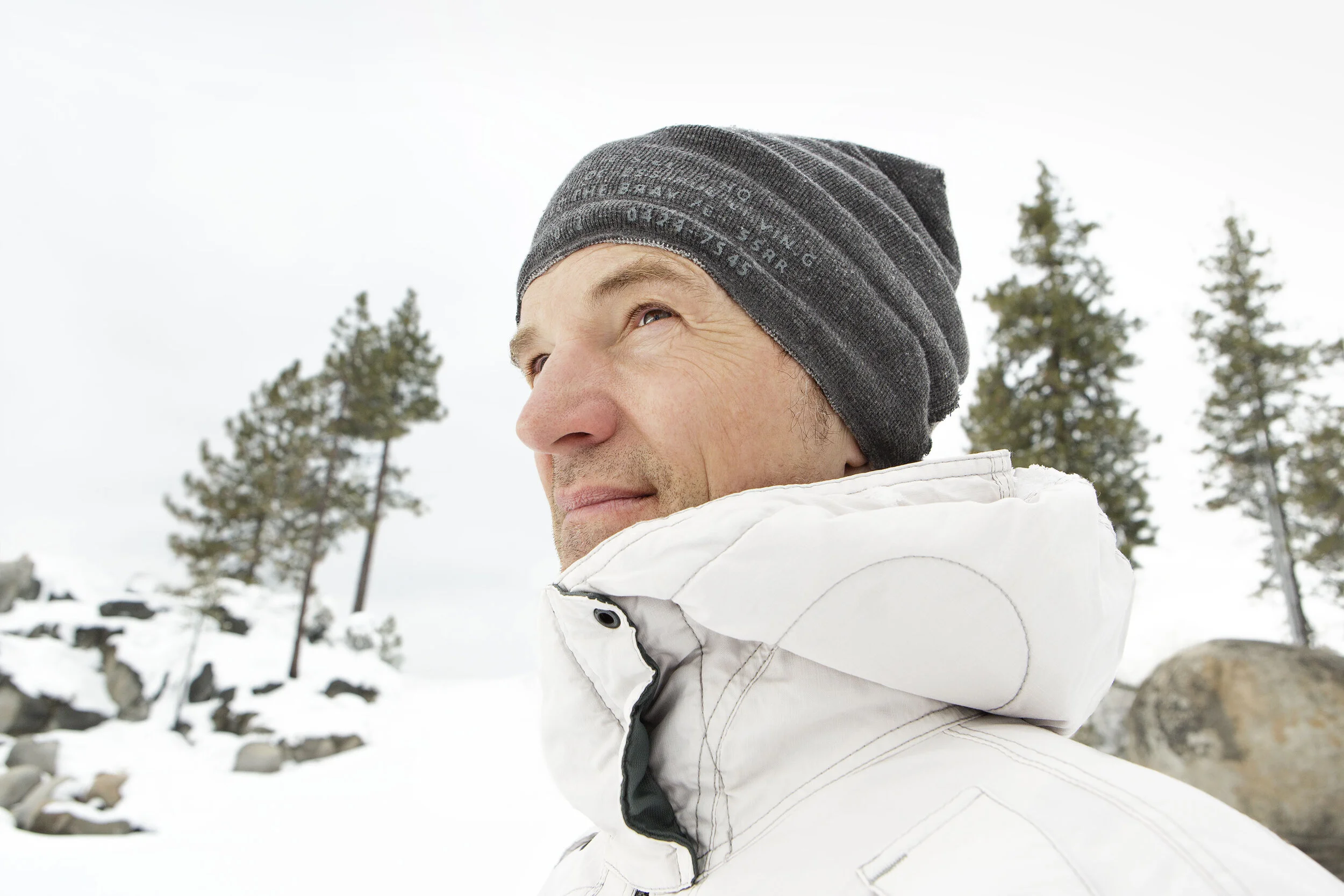 White middle aged man wearing a gray beanie and white winter jacket outdoors in a snowy landscape with trees and rocks at Lake Tahoe, CA.
