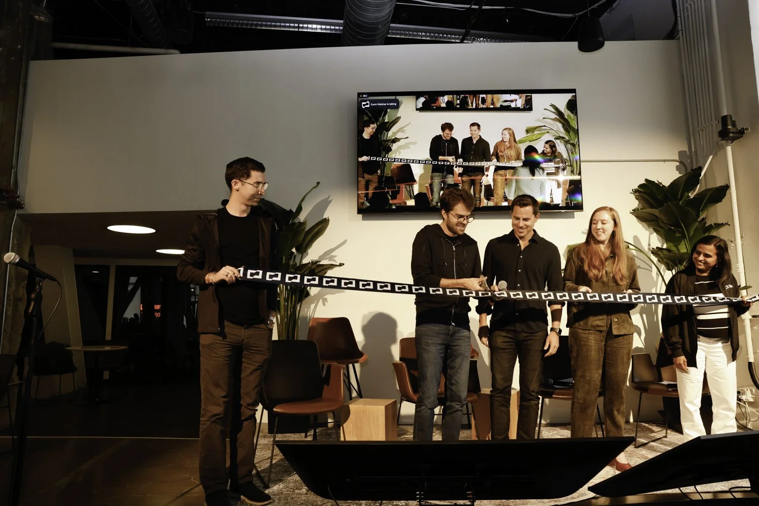 Six people standing on a stage with a microphone, in front of a large screen displaying a group photo, during an event or presentation, with plants and chairs behind them Brex headquarter in San Francisco, CA.