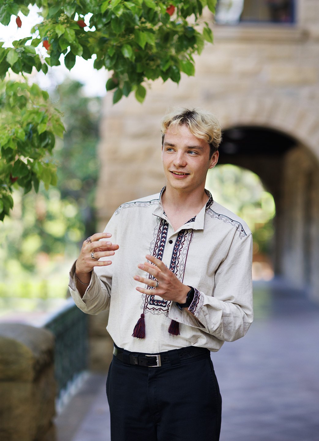 A Ukrainian student at Stanford University with short hair is standing outdoors with a brick arch behind him, gesturing with his hands, with  trees and greenery in the background at Stanford University Campus, Stanford, CA.