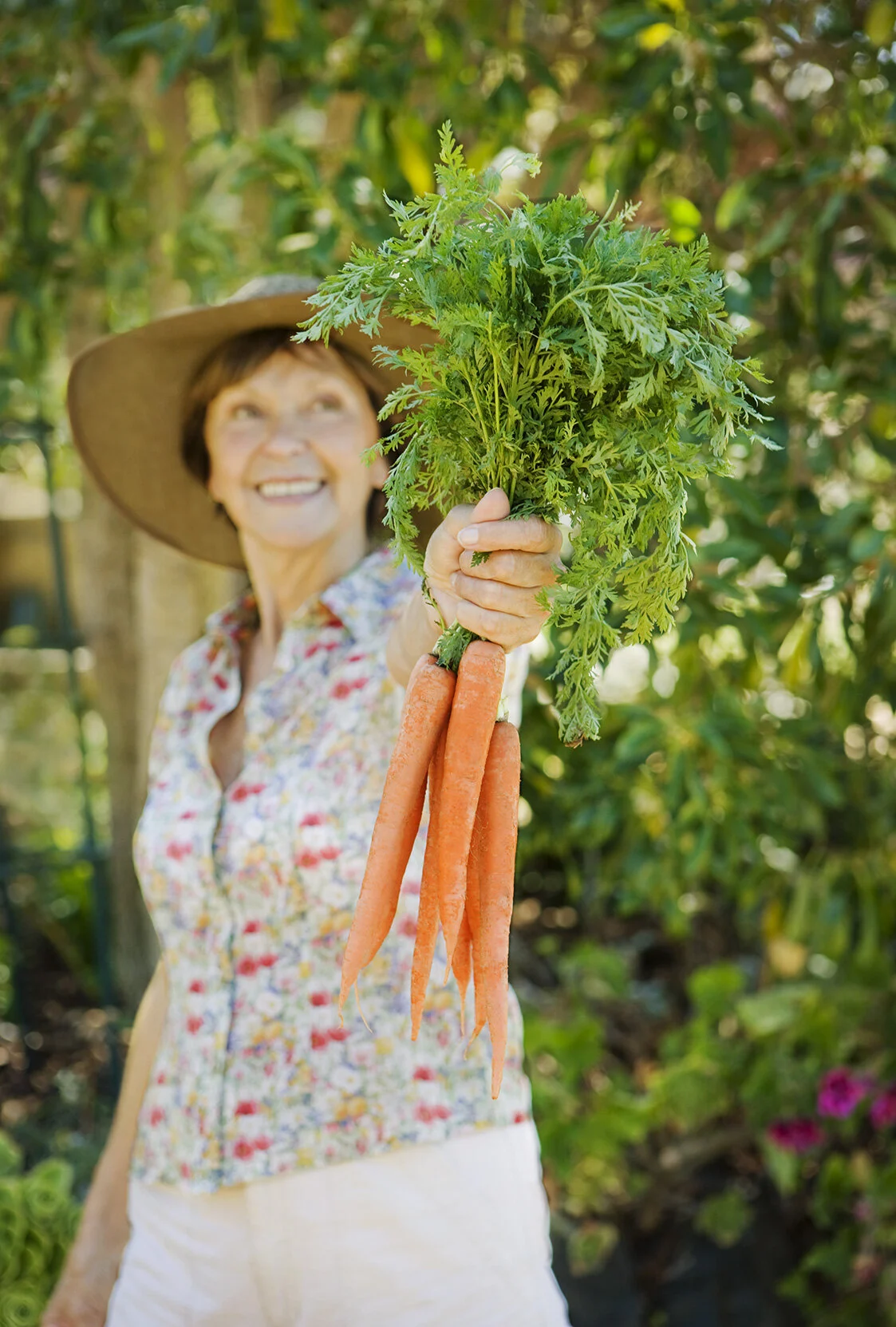 A smiling white middle aged woman wearing a wide-brimmed hat and a floral shirt holding a bunch of fresh carrots with green tops in her outstretched hand in a community garden with flowers and vegetables in San Francisco, CA.