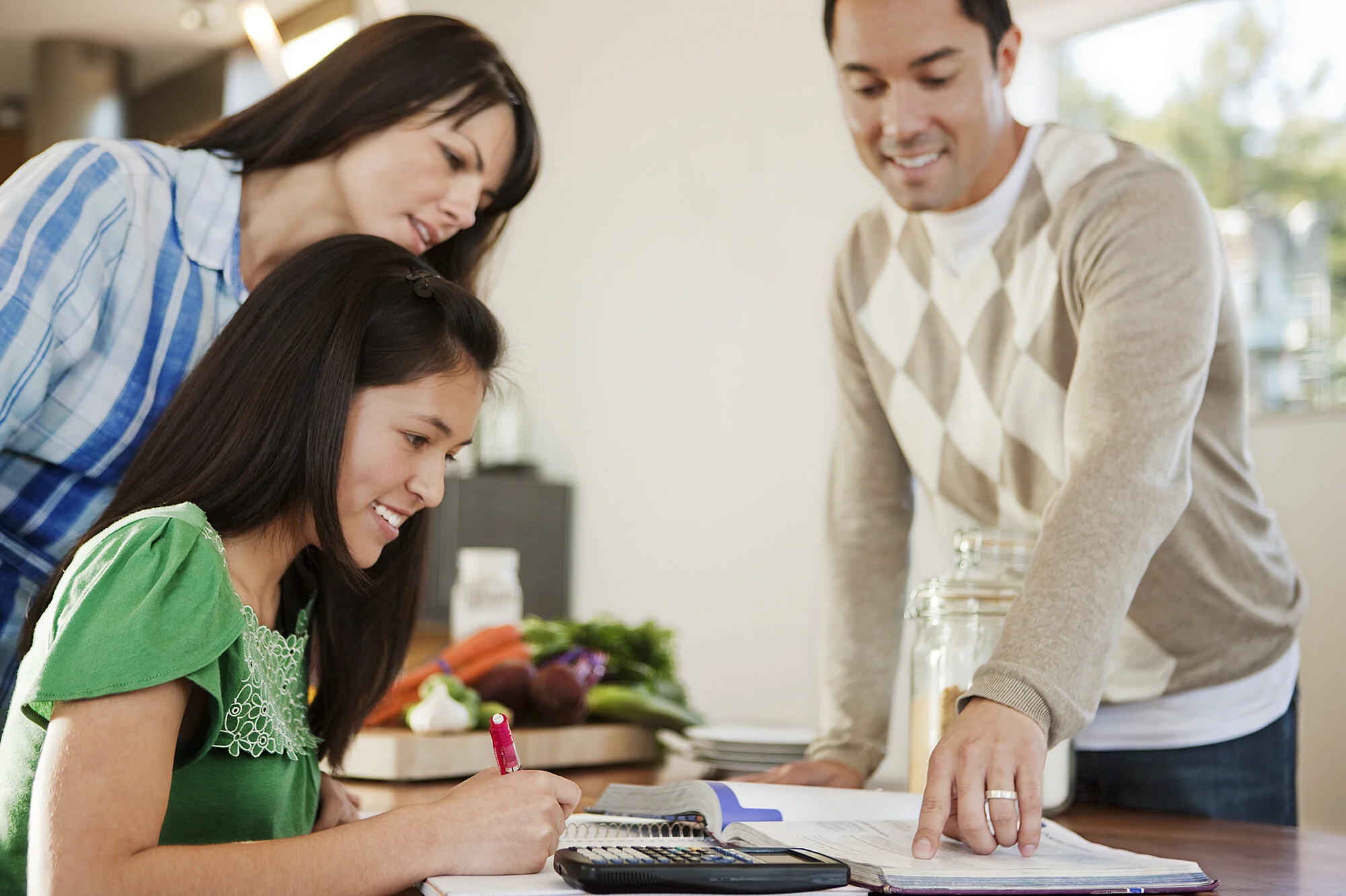 A family standing around a table, smiling and looking at a young girl who is writing and doing homework, with ingredients and vegetables in the background in San Francisco, CA.
