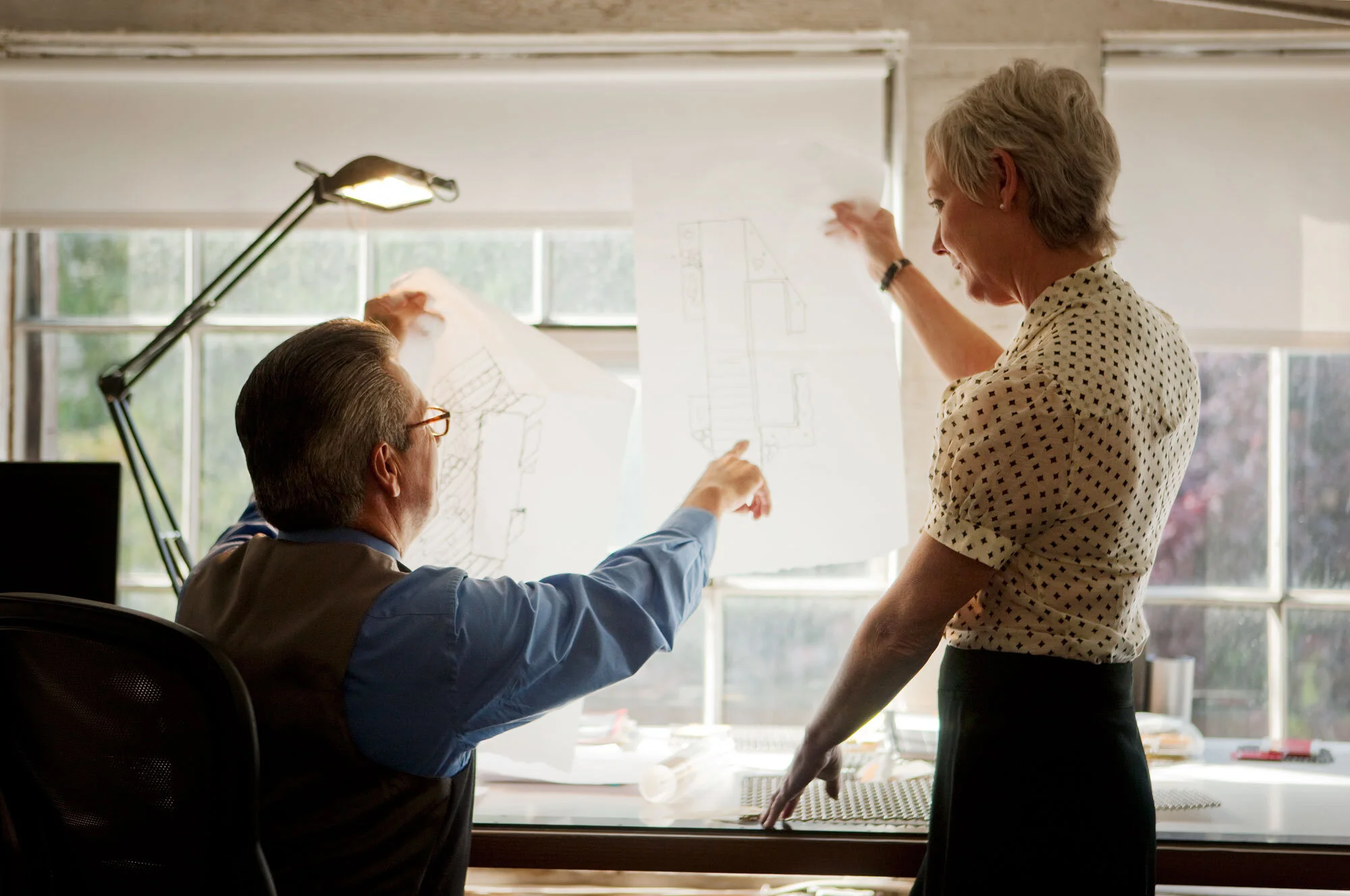 Two professionals discussing architectural blueprints in an office with large windows, natural light, and a desk at an architectural office in San Francisco, CA.