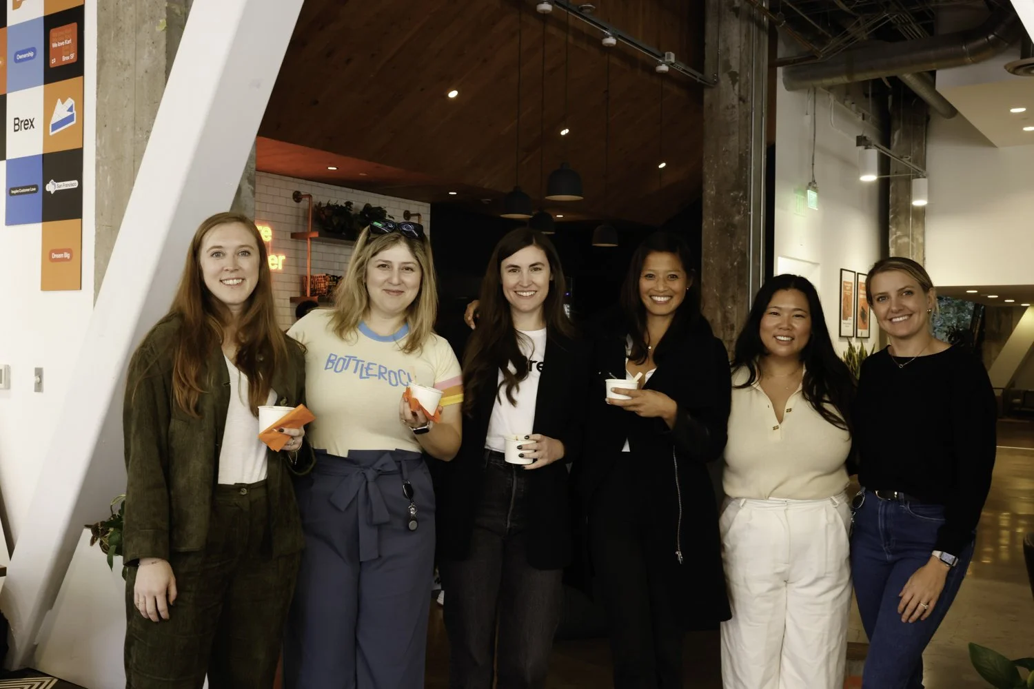 Six women standing together in a modern indoor setting, smiling and holding cups, with a background featuring a mix of brick and industrial-style decor Brex headquarter in San Francisco, CA.