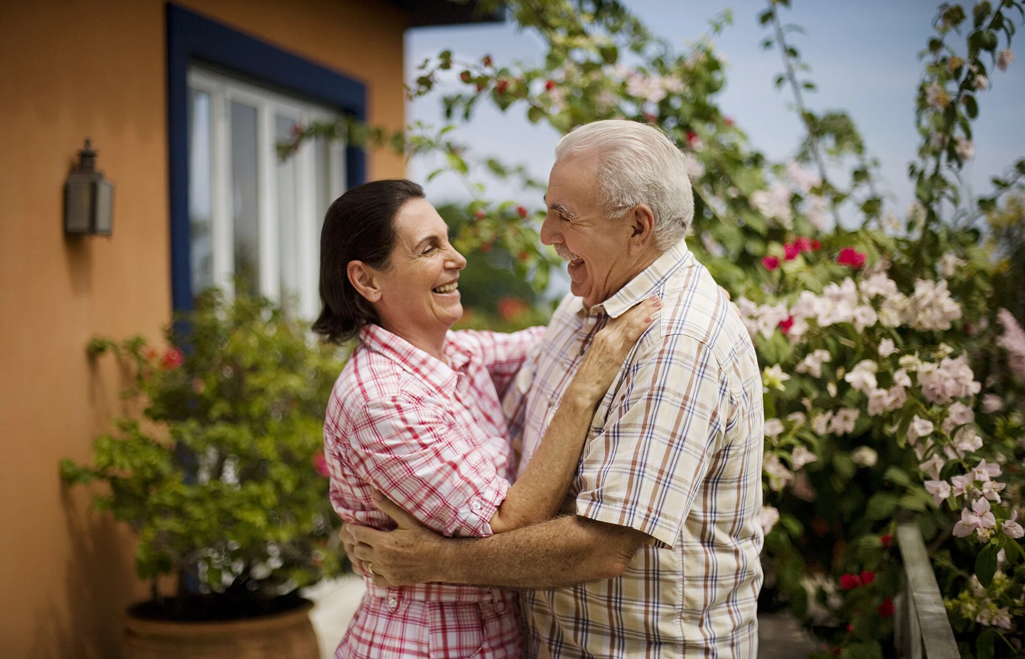 An elderly Brazilian man and woman smiling and embracing each other outdoors and dancing, surrounded by colorful flowers and greenery in Rio de Janeiro, Brazil.