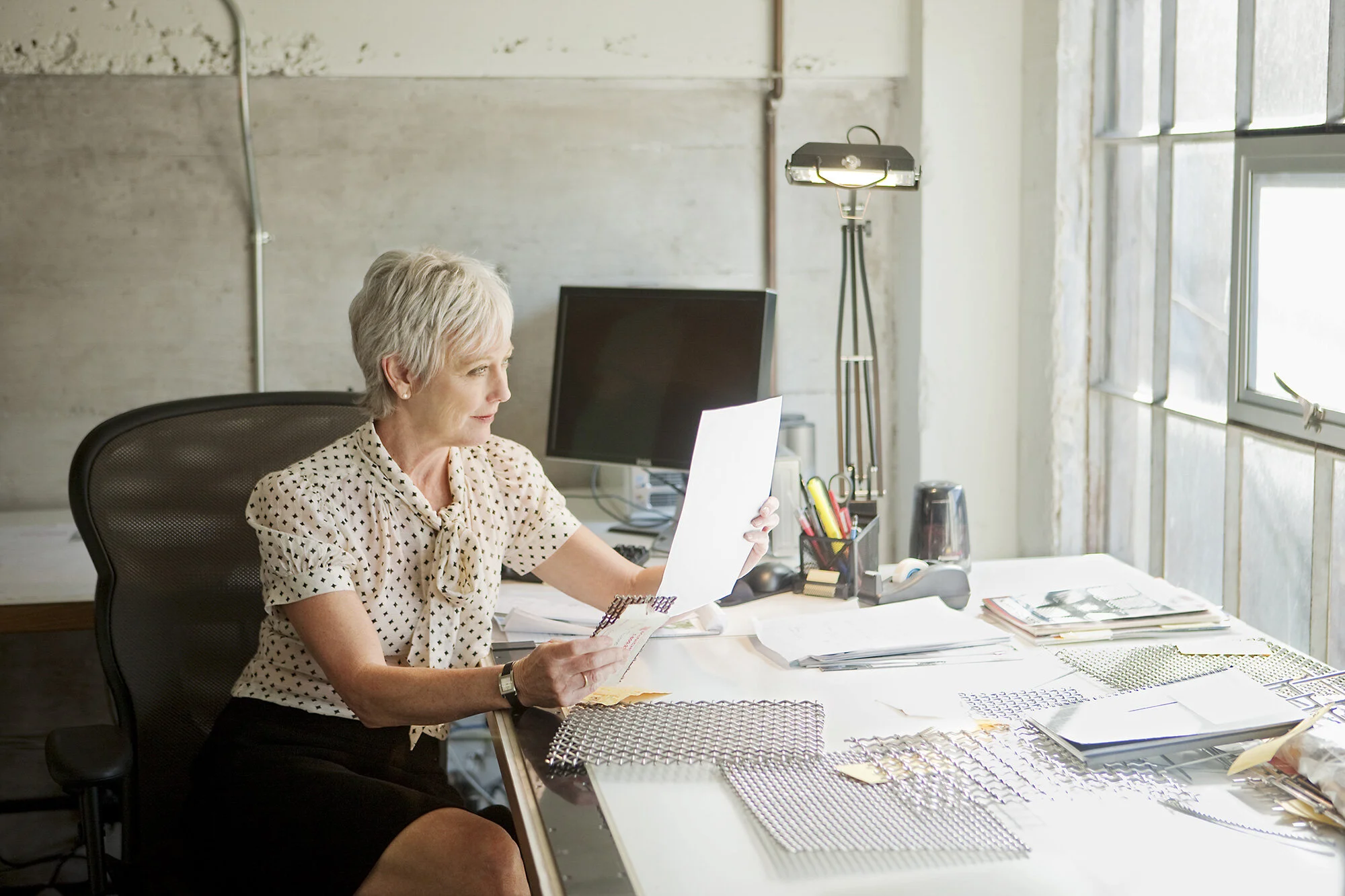 A middle-aged woman with short gray hair sitting at an office desk, looking at a piece of paper. The desk is cluttered with papers, files, and office supplies, and there is a large window to the right providing natural light at an architectural offic