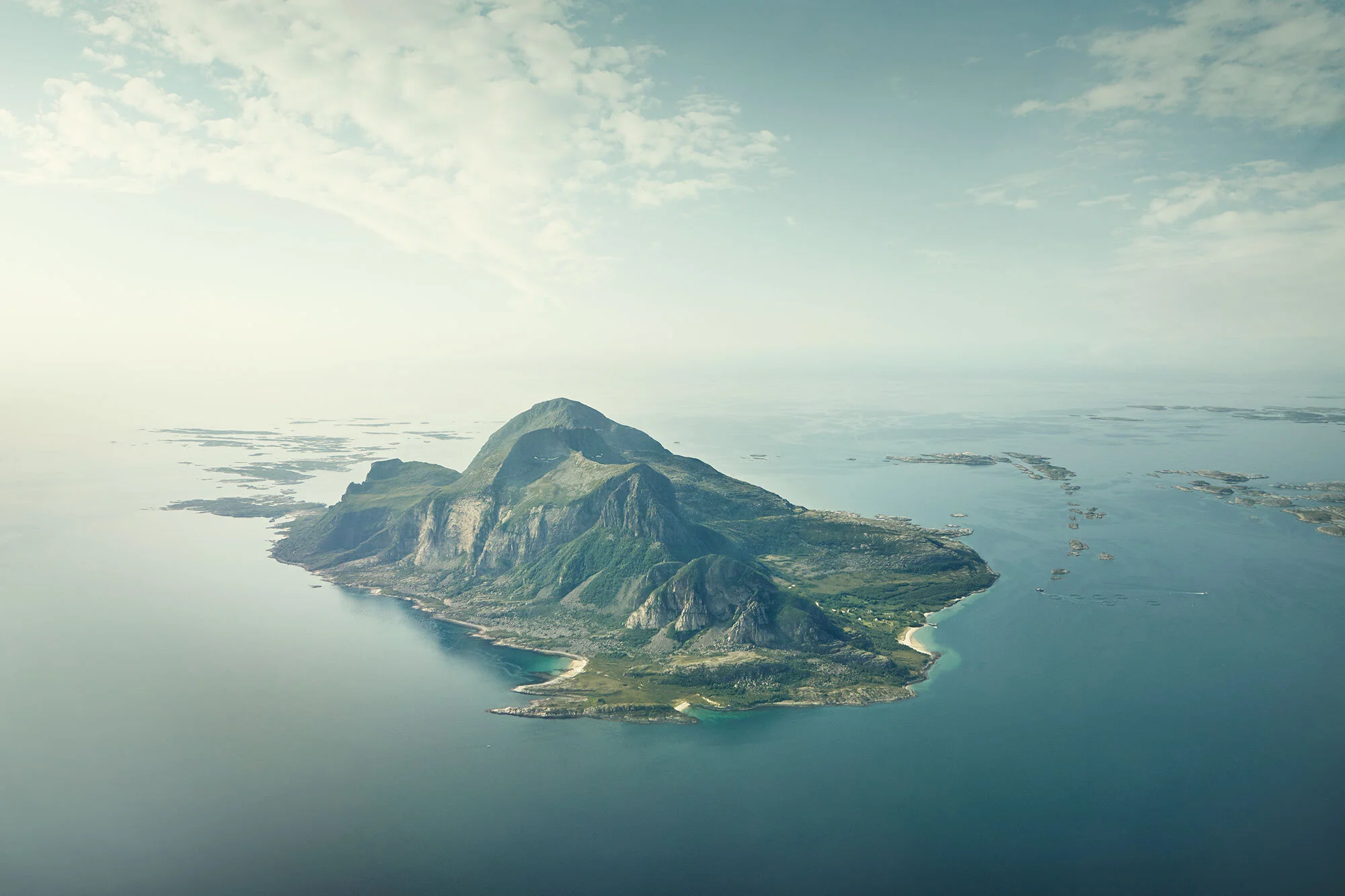 Aerial view of a large, mountainous island surrounded by blue water with small islands nearby, under a partly cloudy sky at the  west coast of Helgelandskysten, Norway.