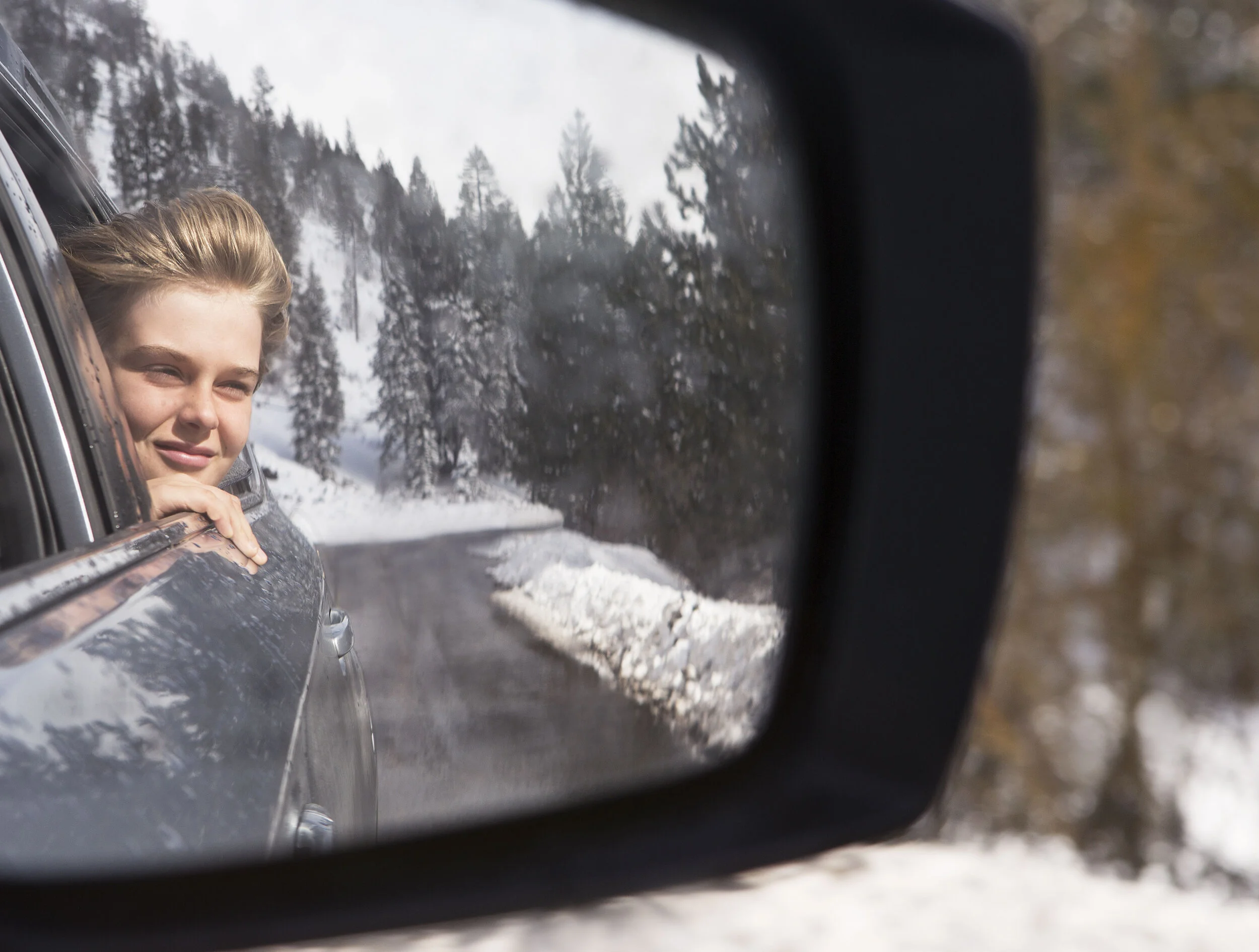 A young white boy looking out of a car window at a snowy mountain landscape, reflected in a side mirror at Lake Tahoe, CA.