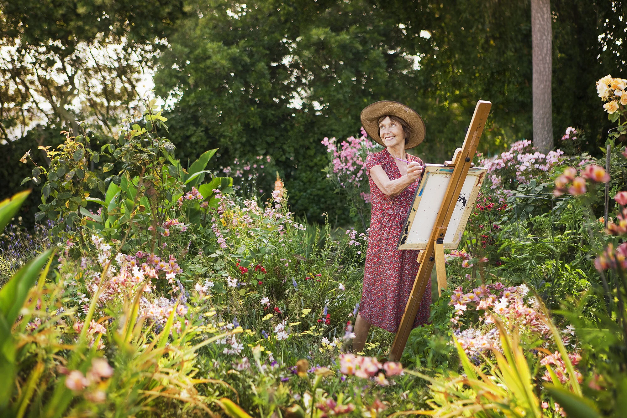 An elderly white woman wearing a red floral dress and a wide-brimmed hat paints on an easel in a lush community garden filled with colorful flowers and green foliage in San Francisco, CA.