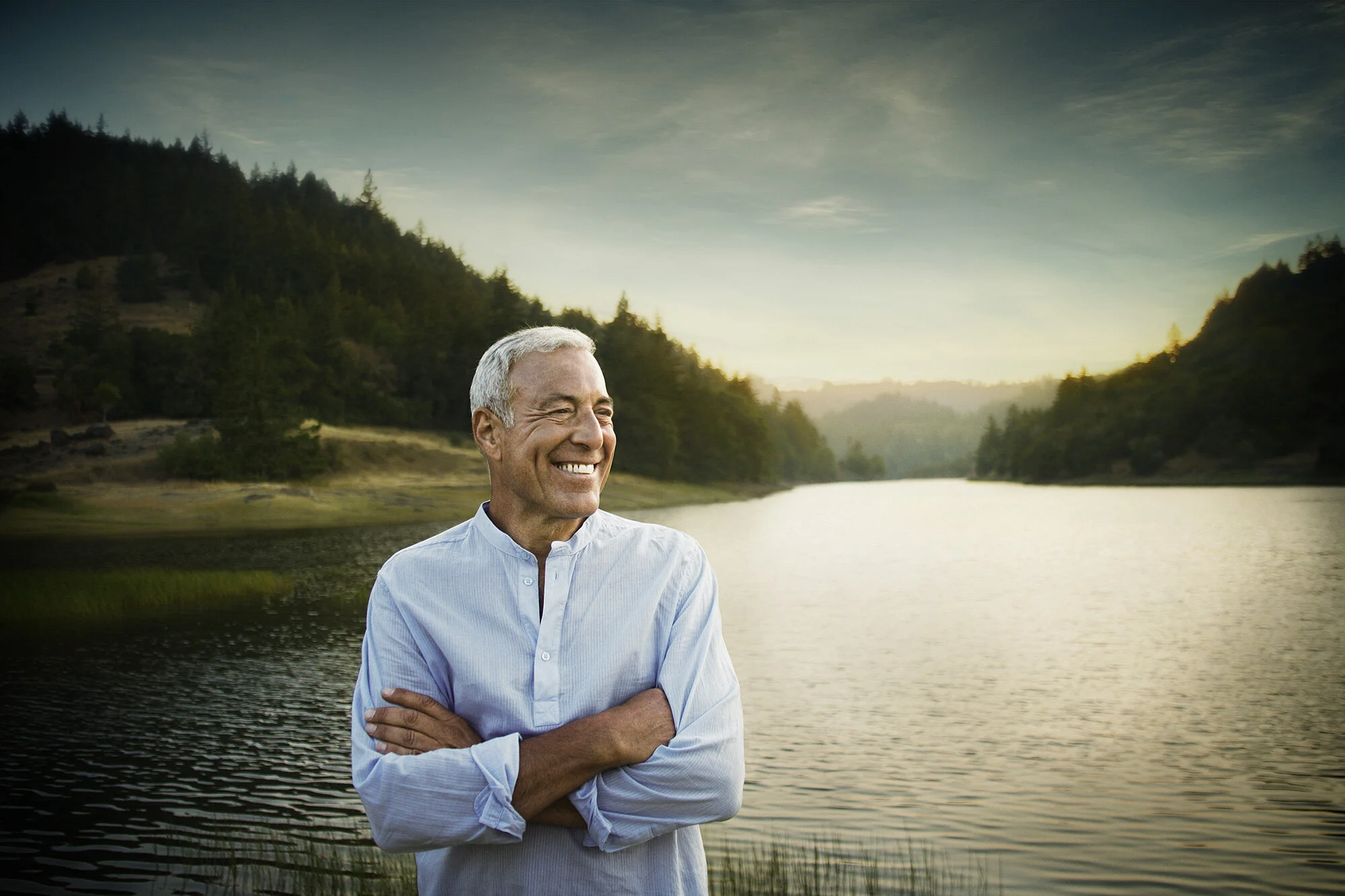 A middle-aged white man with gray hair and a blue shirt, smiling, standing near a lake with trees and hills at sunset in Marin, CA.