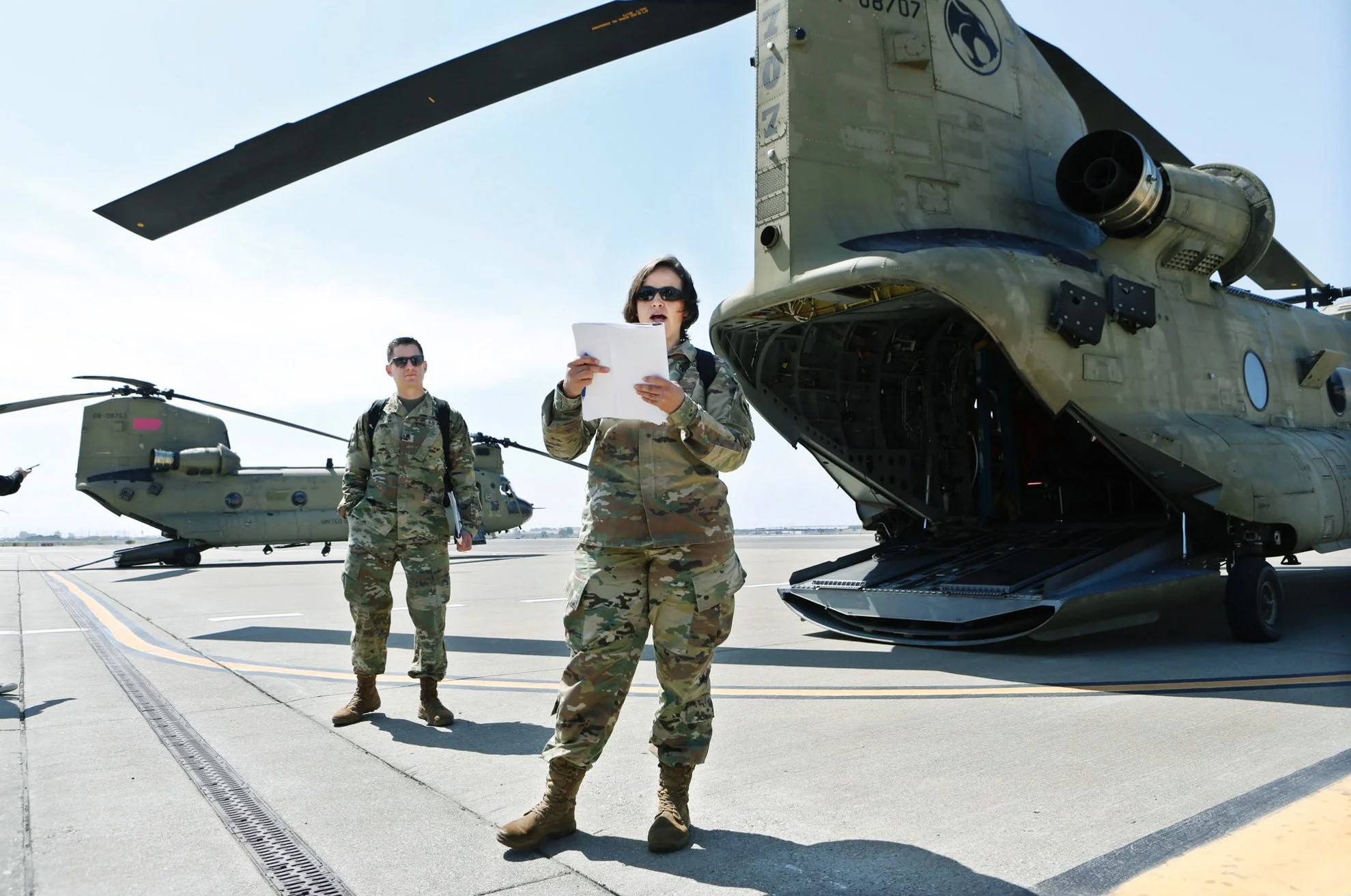 Military personnel reading from a paper in front  of a large military aircraft on an airfield during a clear day. One person is standing on the side of the aircraft in camouflage. Photographed  Salesforce Trailhead Military (Military Veteran Recruitm