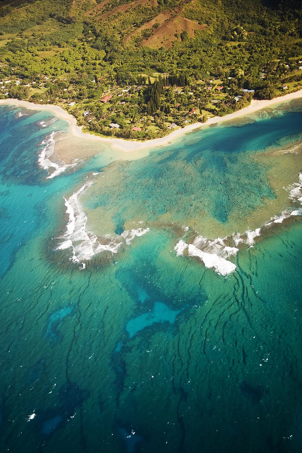 Aerial view of a coastal area with lush green hills, a sandy beach, and turquoise ocean water with visible coral reefs and waves in Kaua'i, Hawai'i.