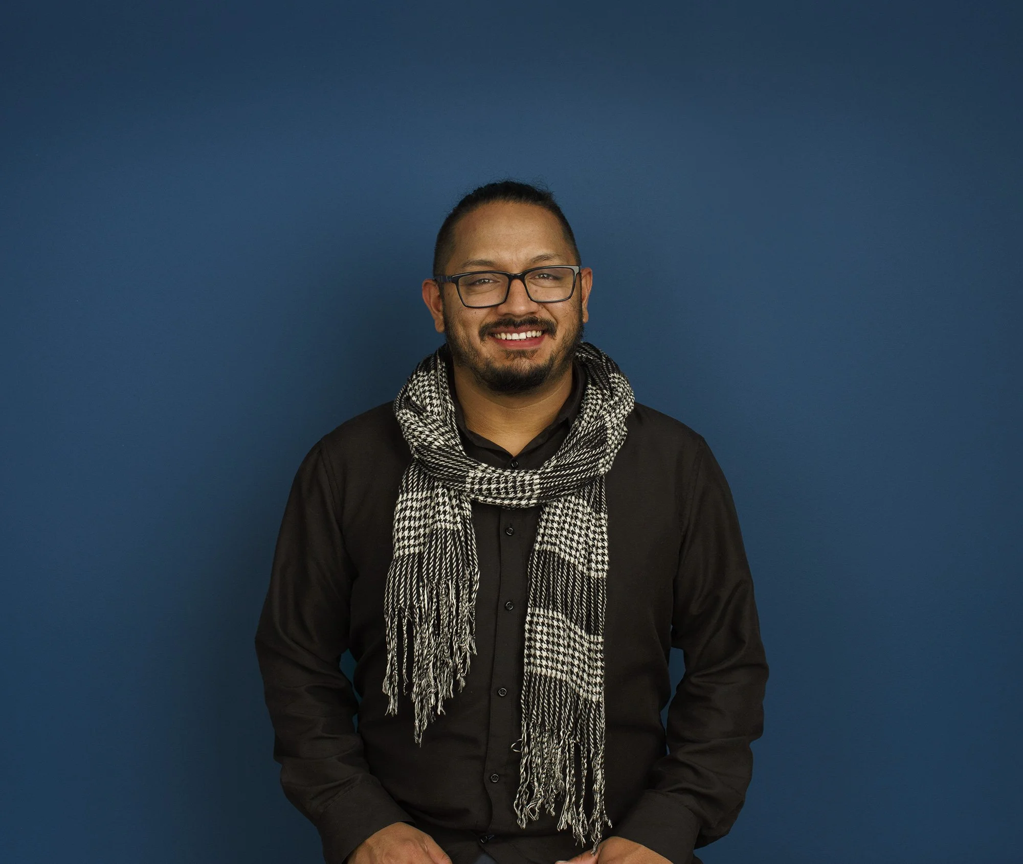 Latino man in black shirt and glasses standing against a blue background, smiling, with a black and white scarf around his neck at an architectural office in San Francisco, CA.