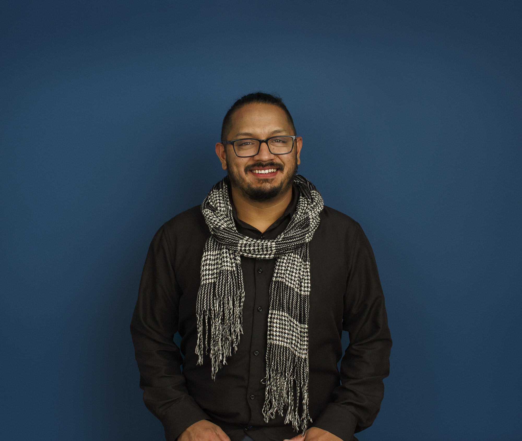 Latino man in black shirt and glasses standing against a blue background, smiling, with a black and white scarf around his neck at an architectural office in San Francisco, CA.