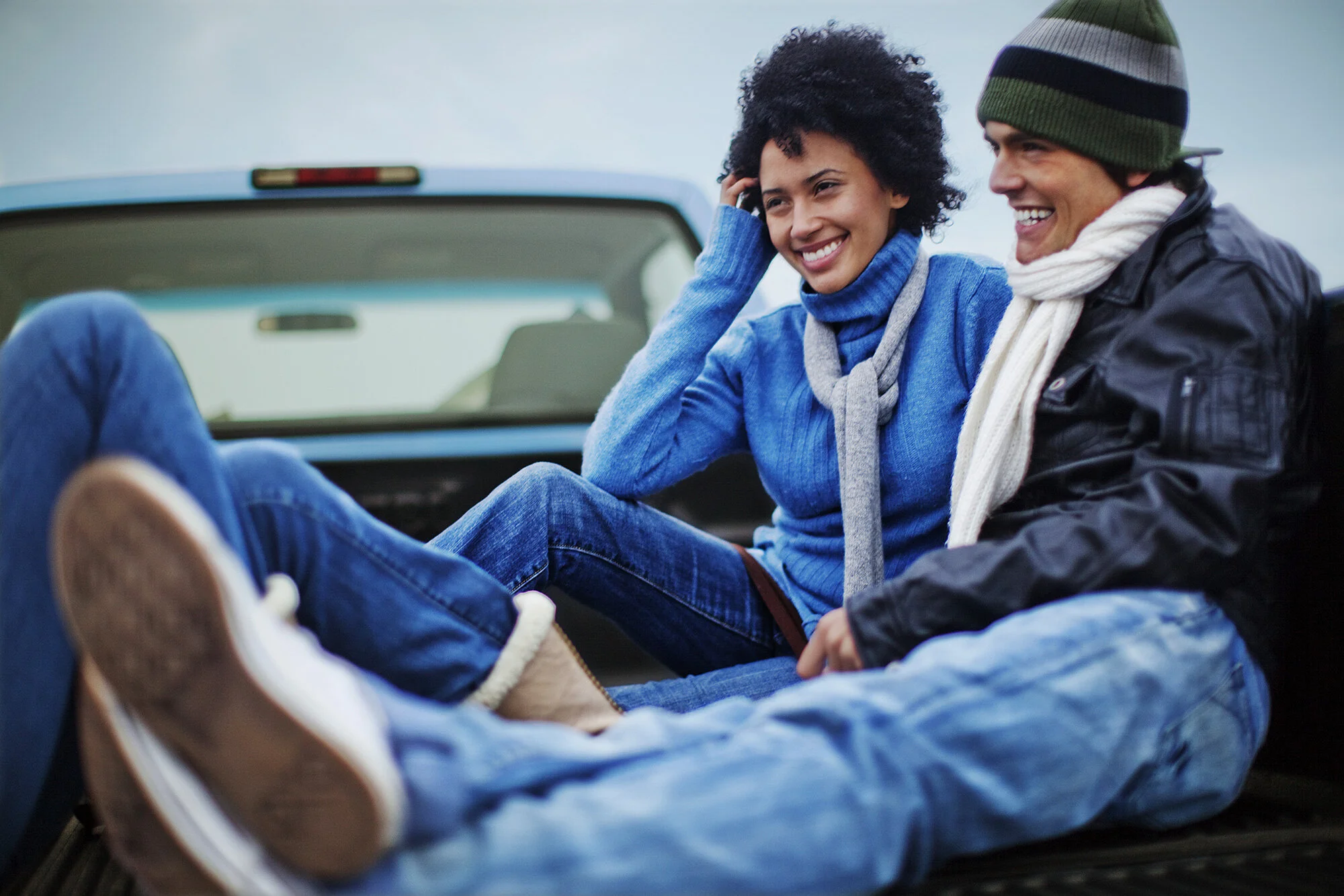 A couple, a Black young woman and Latino young man, smiling people sitting in the back of a pickup truck, dressed in casual and warm clothing, outdoors on a cloudy day at Ocean Beach in San Francisco, CA.