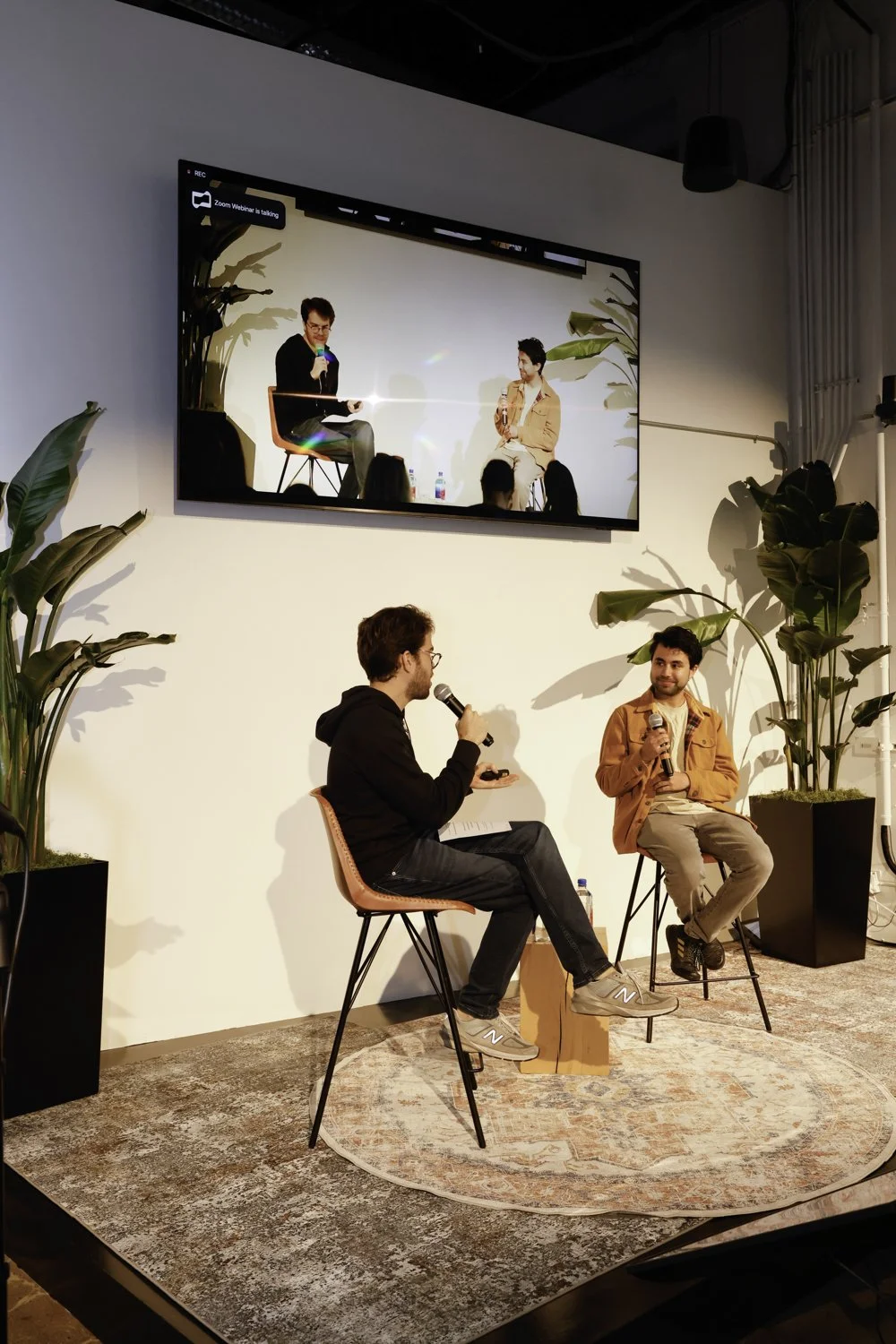 Two men are sitting on chairs during a panel discussion or interview, with one holding a microphone. A large screen above them displays the same scene. The setting includes potted plants and a textured rug on the floor Brex headquarter in San Francis