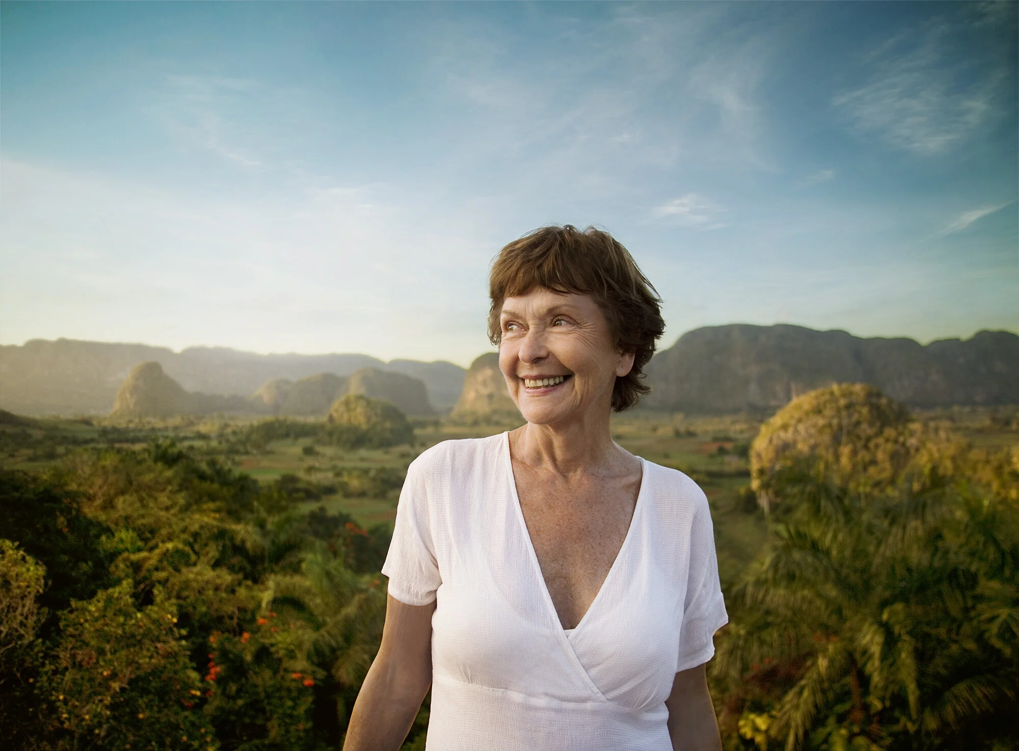 Smiling middle aged woman with short brown hair outdoors in a lush, green landscape with mountains in the background under a blue sky in Vinales, Cuba.