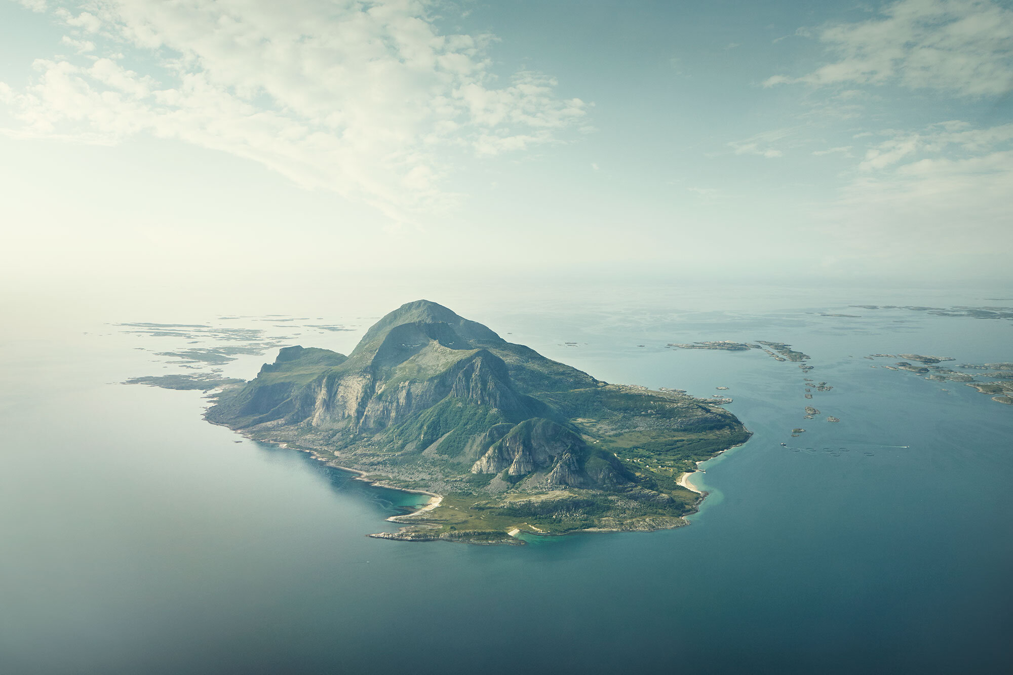 Aerial view of a large island with mountainous terrain surrounded by water shaped like an Easter Island statue, with a small beach on the island's shoreline along Norway's western coastline called Helgelandskysten, Norway.