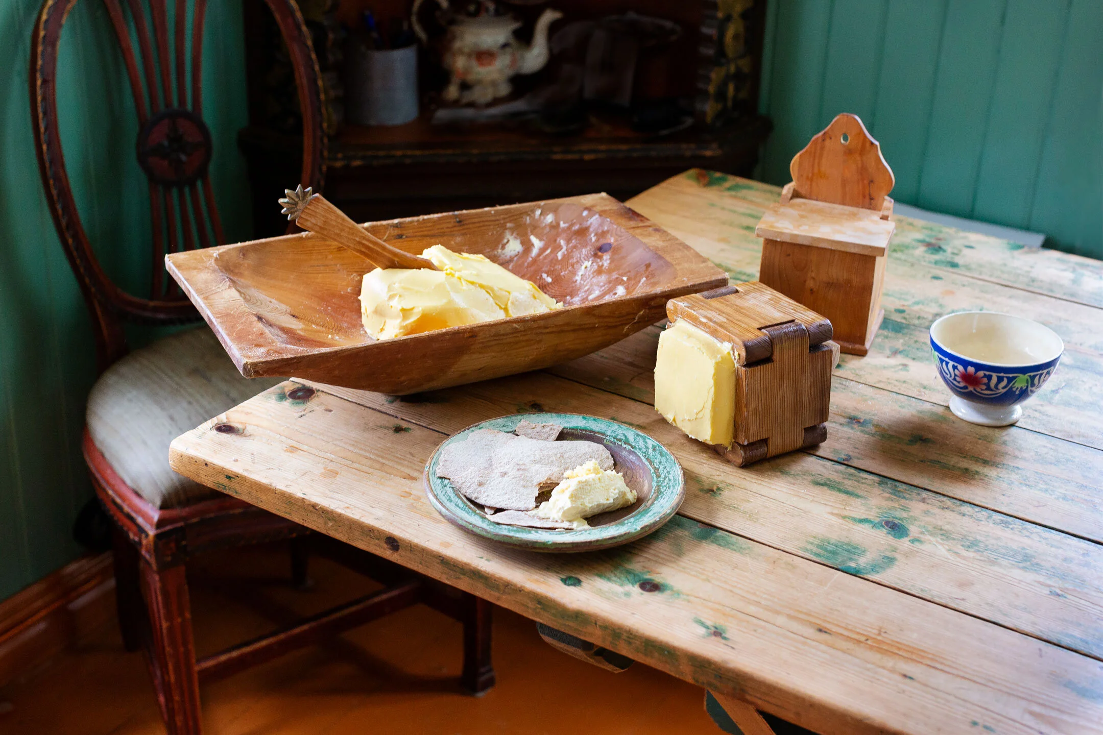 A rustic wooden table with a large wooden bowl containing butter, a small wooden butter mold, a dish with bread and butter, a decorative bowl with spices, and a small wooden box with a lid. The background features a wooden chair and a cabinet in Fann