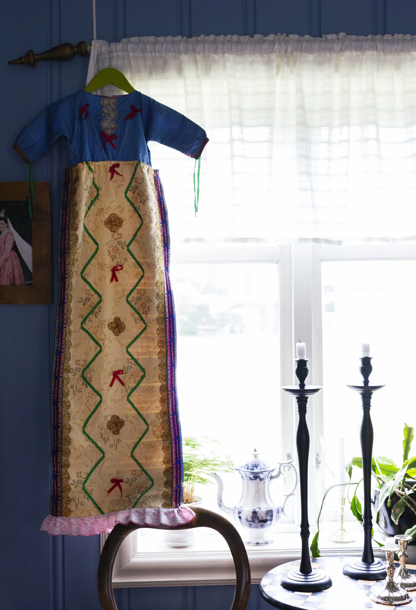 Colorful decorative Norwegian baptism dress hanging on curtain rod in a window, with candle holders, teapot, and plants on a table below, in a bright room with white curtains in Fannrem, Norway.