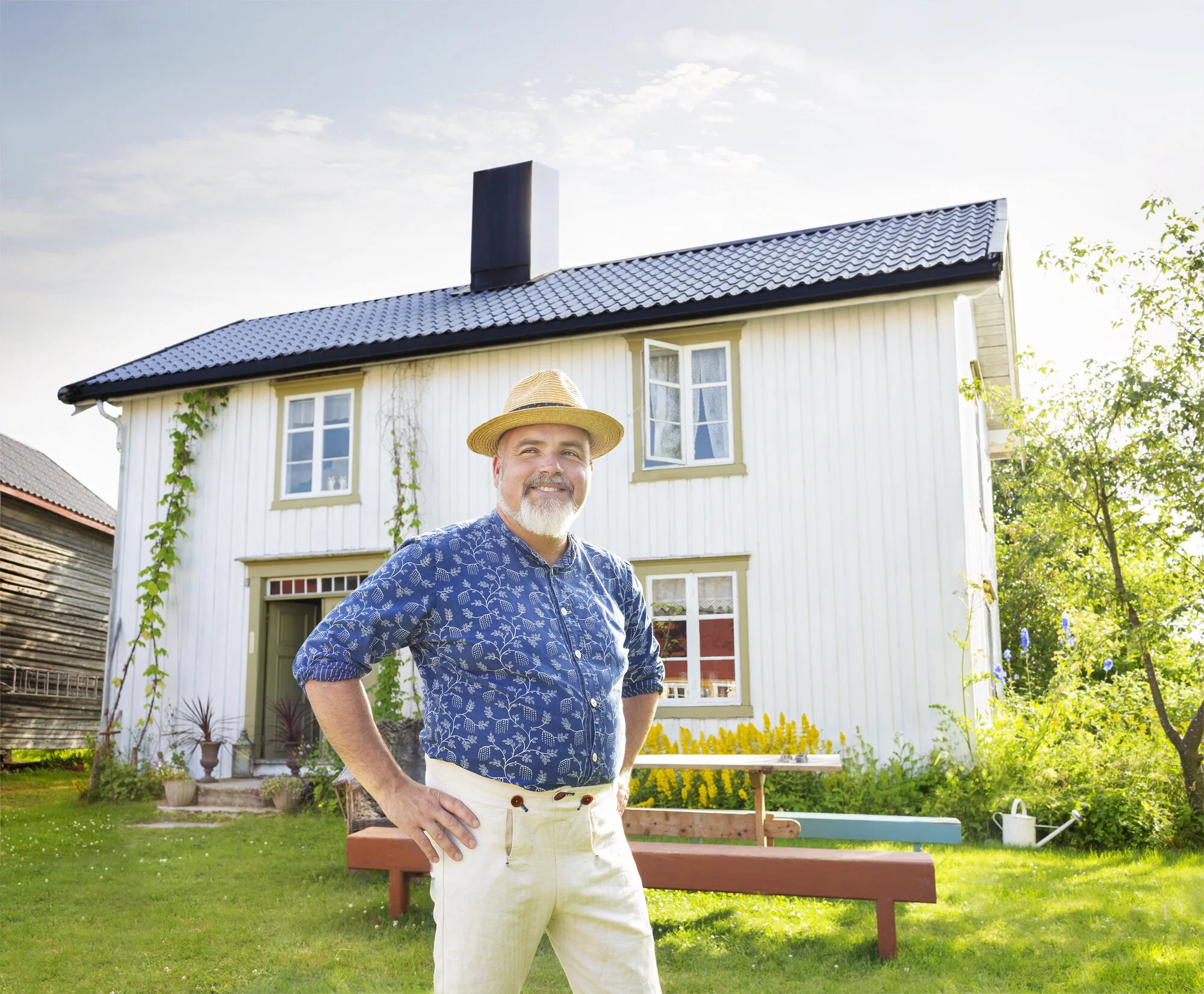 A smiling Norwegian farmer  with a gray beard wearing a straw hat, a blue patterned shirt, and white pants, standing outdoors in front of a white two-story farm house with a black roof surrounded by greenery and flowers in Fannrem, Norway.