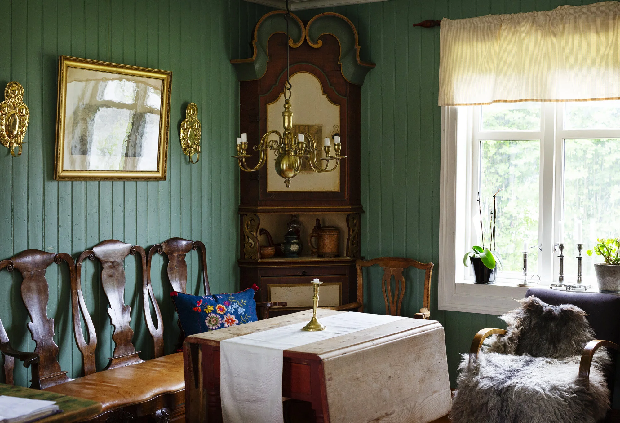 A vintage Norwegian dining room with green wooden panel walls,  chairs, a wooden table with a white linen tablecloth, a brass chandelier, a mirror,  flowers in pots on the windowsill, and a sheepskin-covered armchair by the window in Fannrem, Norway.