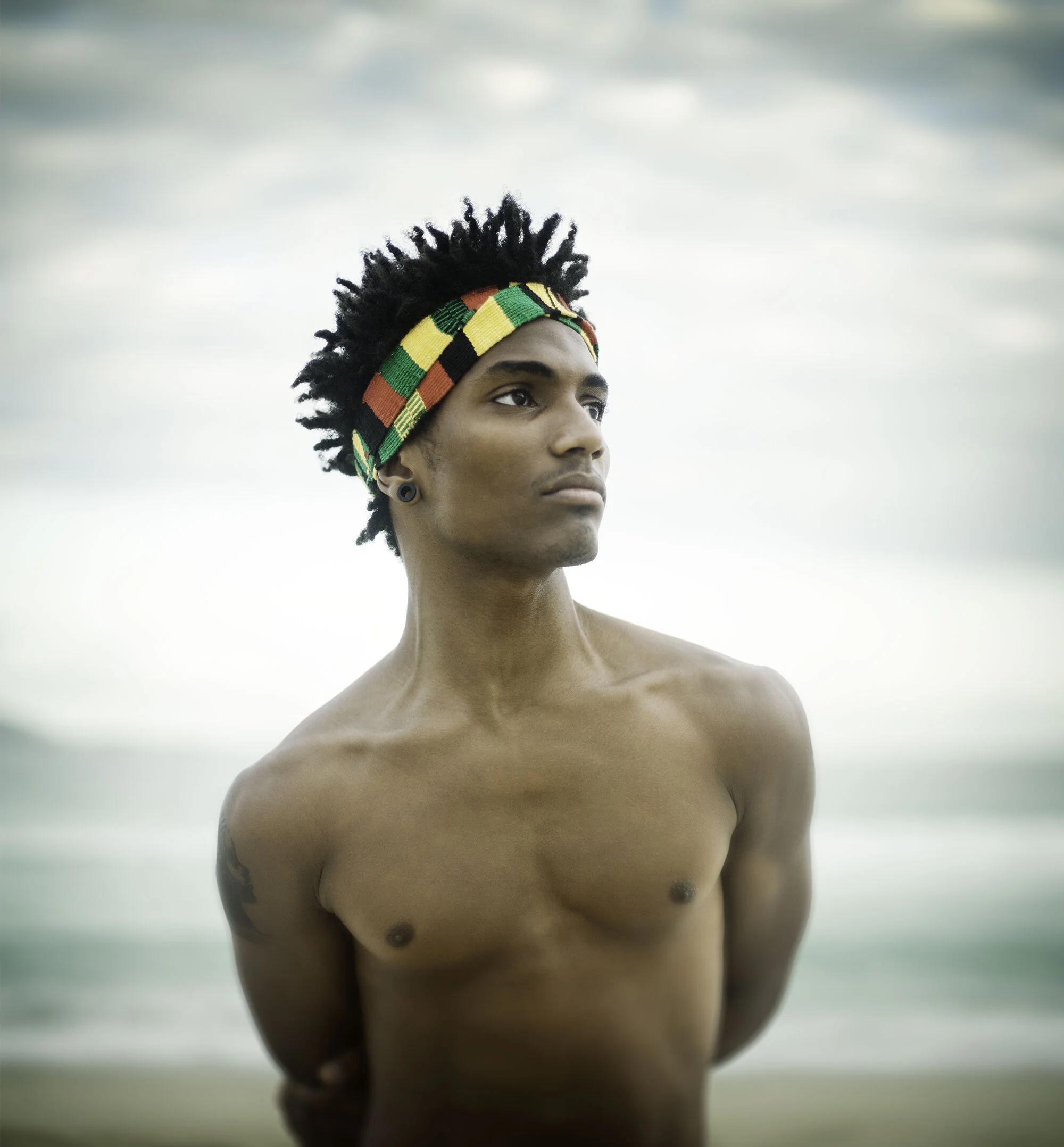 A shirtless, fit, young, Black Brazilian man, black curly hair, and a colorful Rasta headband standing on Ipanema beach with cloudy skies in the background in Rio de Janeiro, Brazil.