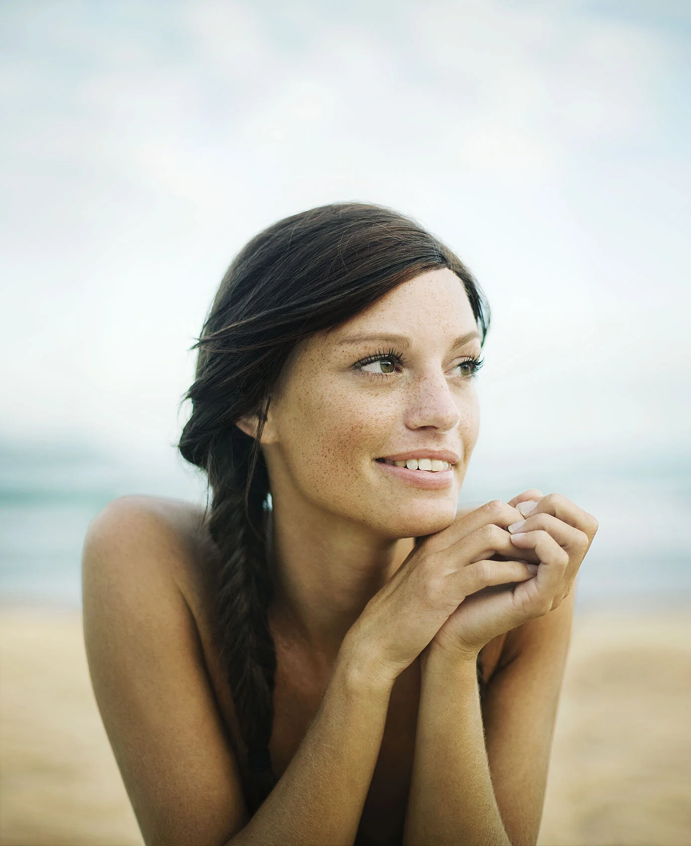 A white woman with dark brown hair in a braid, sitting outdoors at the beach, smiling softly with her hands near her chin, looking to the side in Ipanema beach in Rio de Janeiro, Brazil.