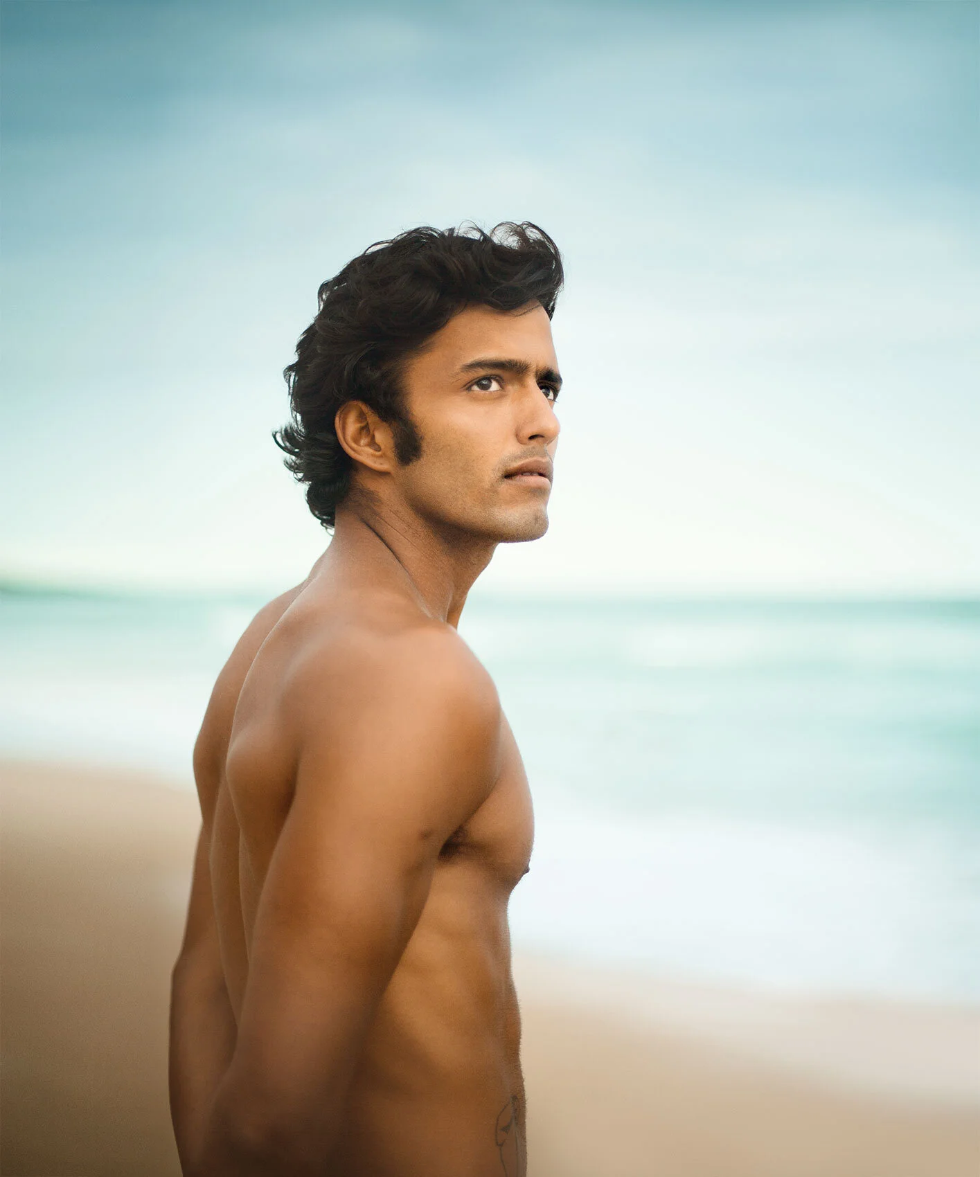 A Brazilian shirtless man with dark wavy black hair standing on Ipanema beach, looking to the side with a contemplative expression, ocean and sky in the background in Rio de Janeiro, Brazil.