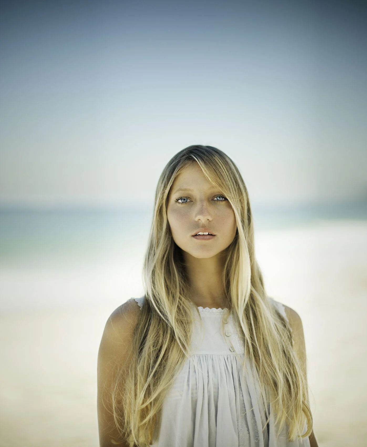 Portrait of a young Brazilian woman with long blonde hair and blue eyes standing on Ipanema beach with blurred ocean in the background, wearing a white dress looking at the camera in Rio de Janeiro, Brazil.