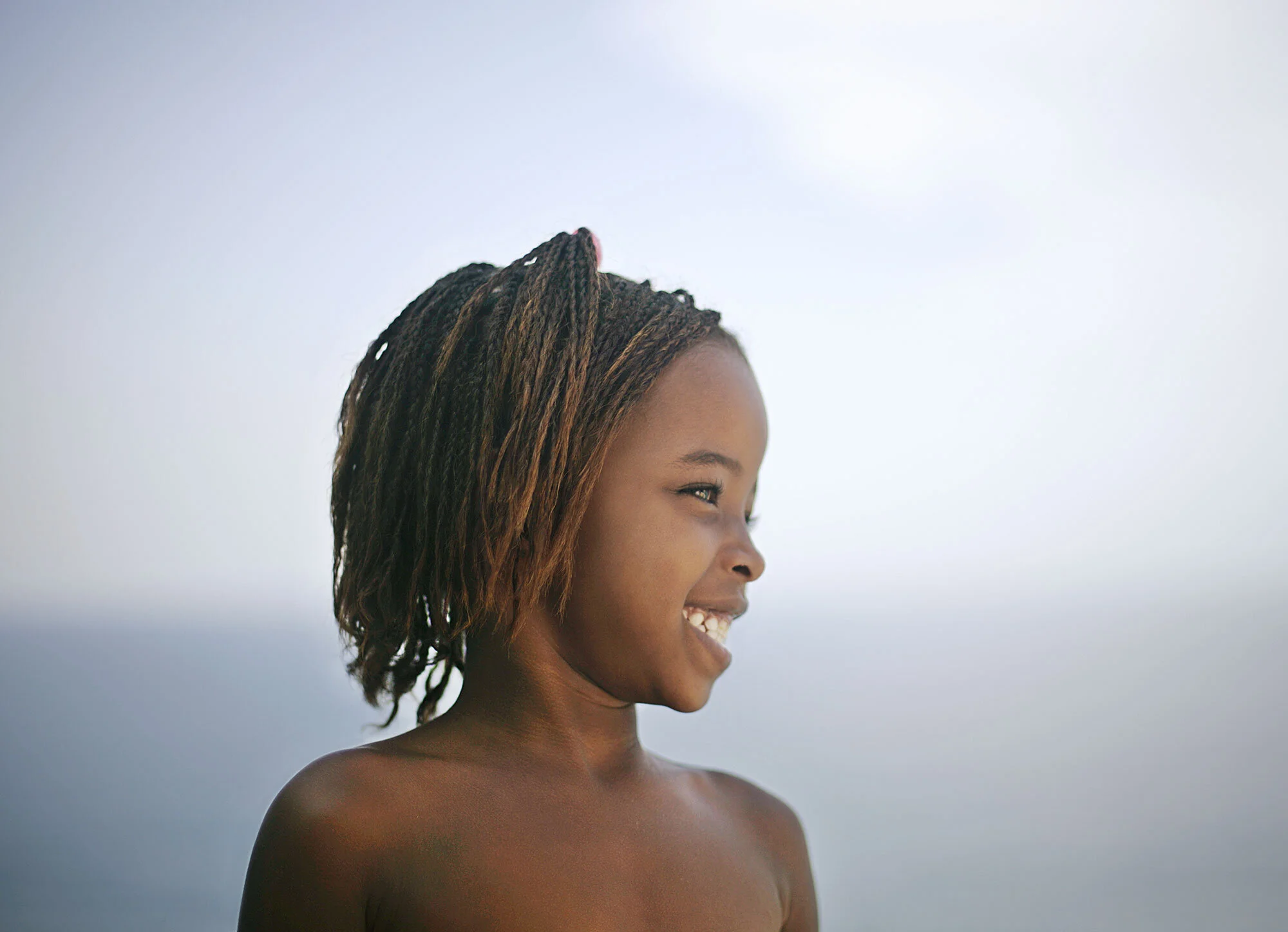 A young Black Brazilian girl with braided hair smiling while facing to the right, with a blurred sky and ocean in the background at Ipanema beach in Rio de Janeiro, Brazil.