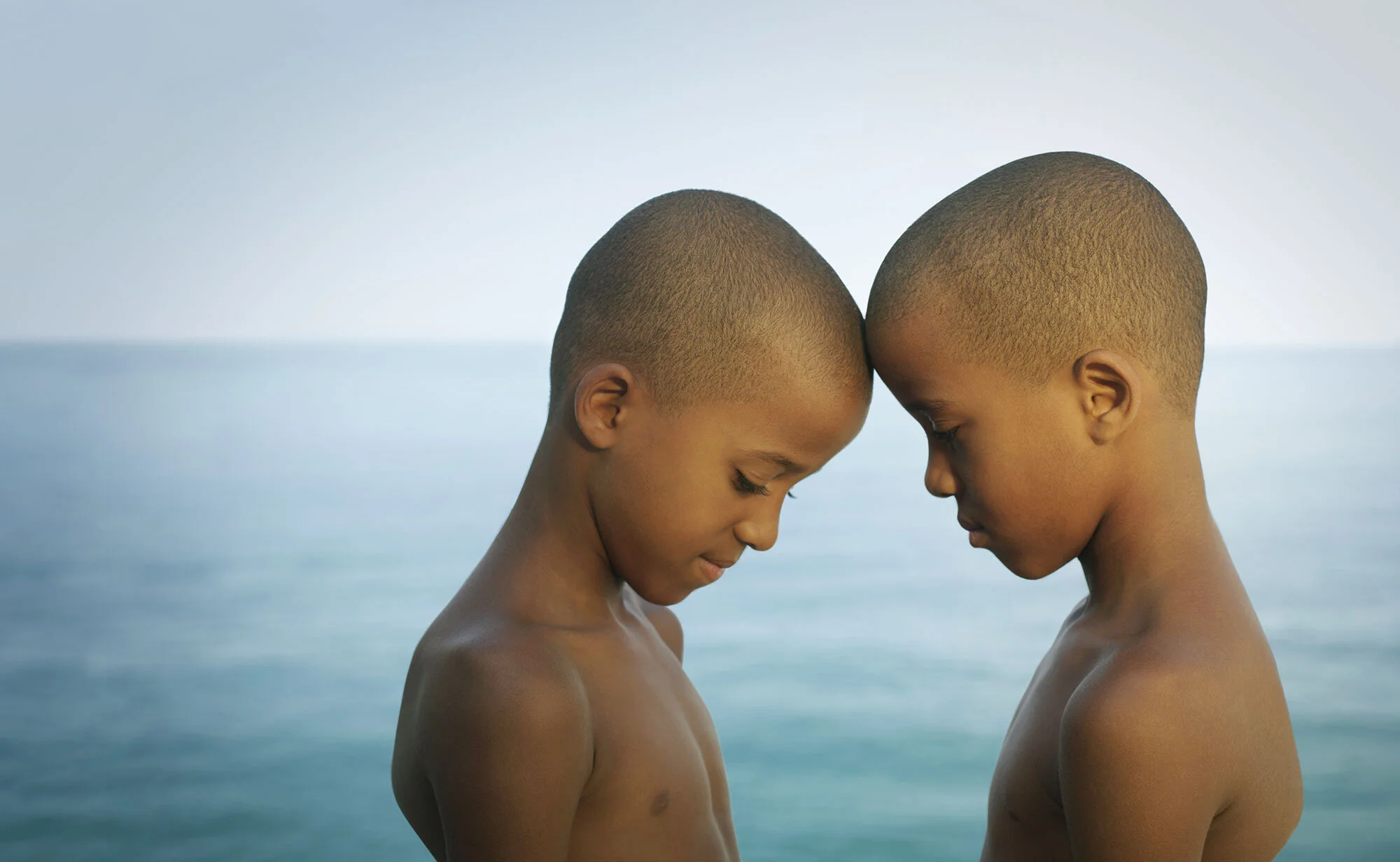 Two young Brazilian Black boys with shaved heads and no shirts are touching foreheads with their eyes closed, standing in front of a calm Atlantic ocean at Ipanema beach in Rio de Janeiro, Brazil.