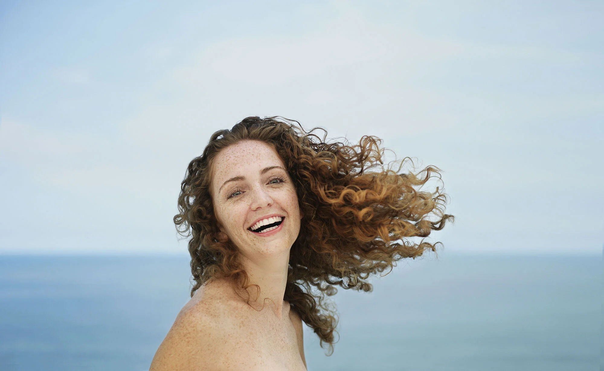 A young, white, Brazilian woman with curly red hair and freckles smiling at the camera, is throwing her hair in the air, set against a beach and ocean background with a blue sky at Ipanema beach, in Rio de Janeiro, Brazil.