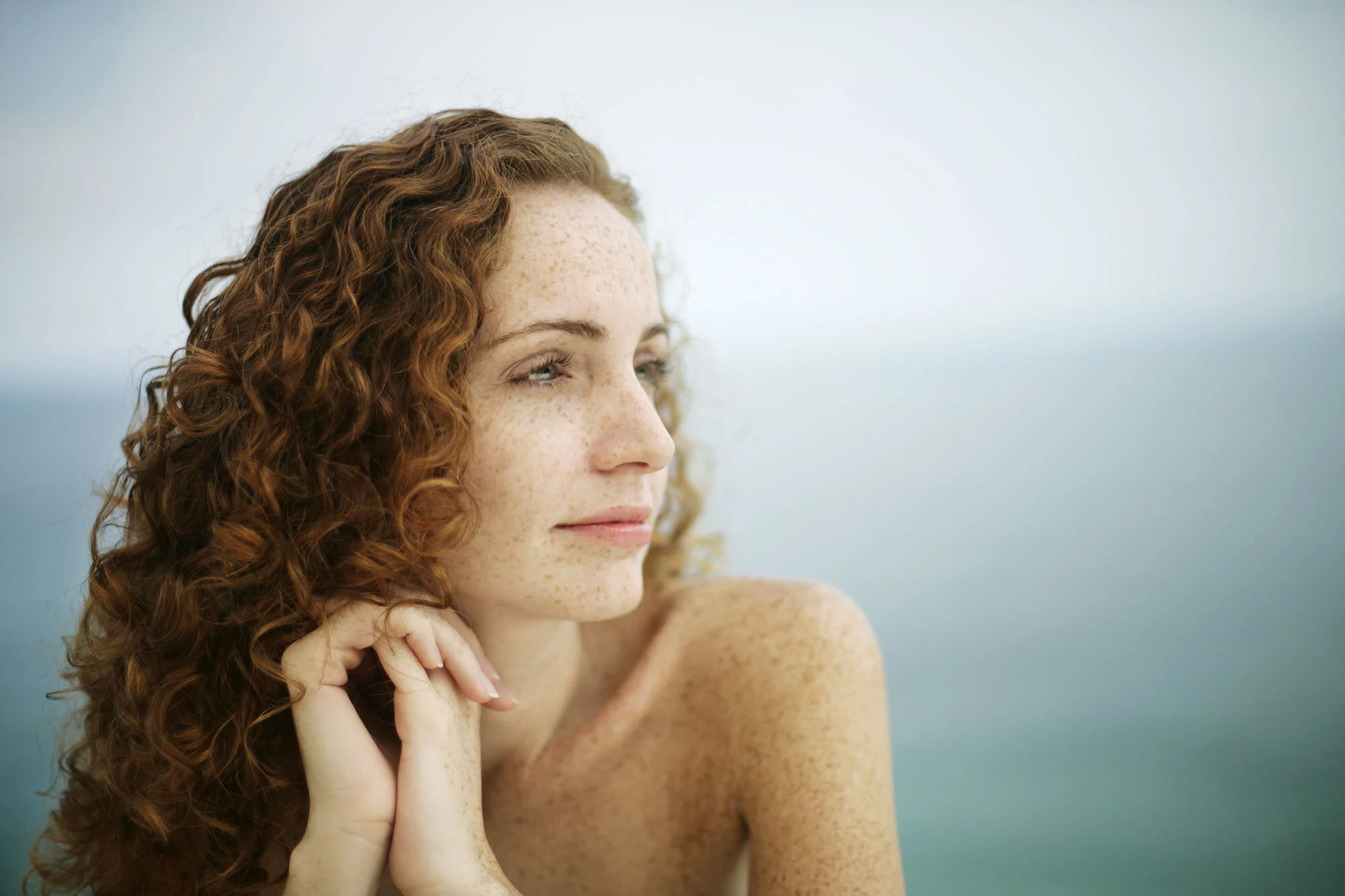 Close-up of a woman with red curly hair and freckles, looking to the side with a serene expression, outdoors with a blurred blue background at Ipanema beach in Rio de Janeiro, Brazil.