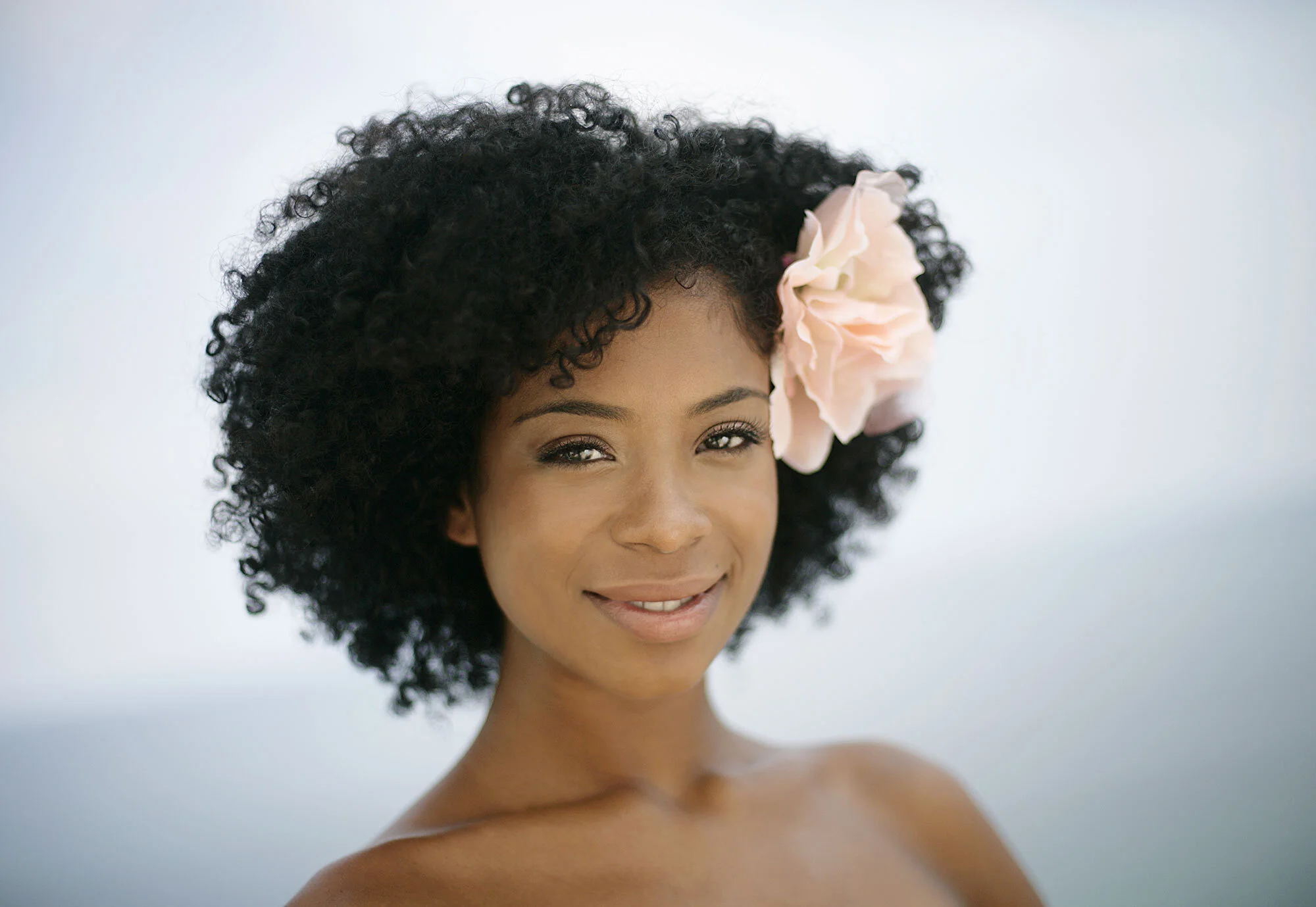 A Black Brazilian woman with dark curly hair and a large pink flower in her hair, smiling softly with bare shoulders, against a soft blue background at Ipanema beach in Rio de Janeiro, Brazil.