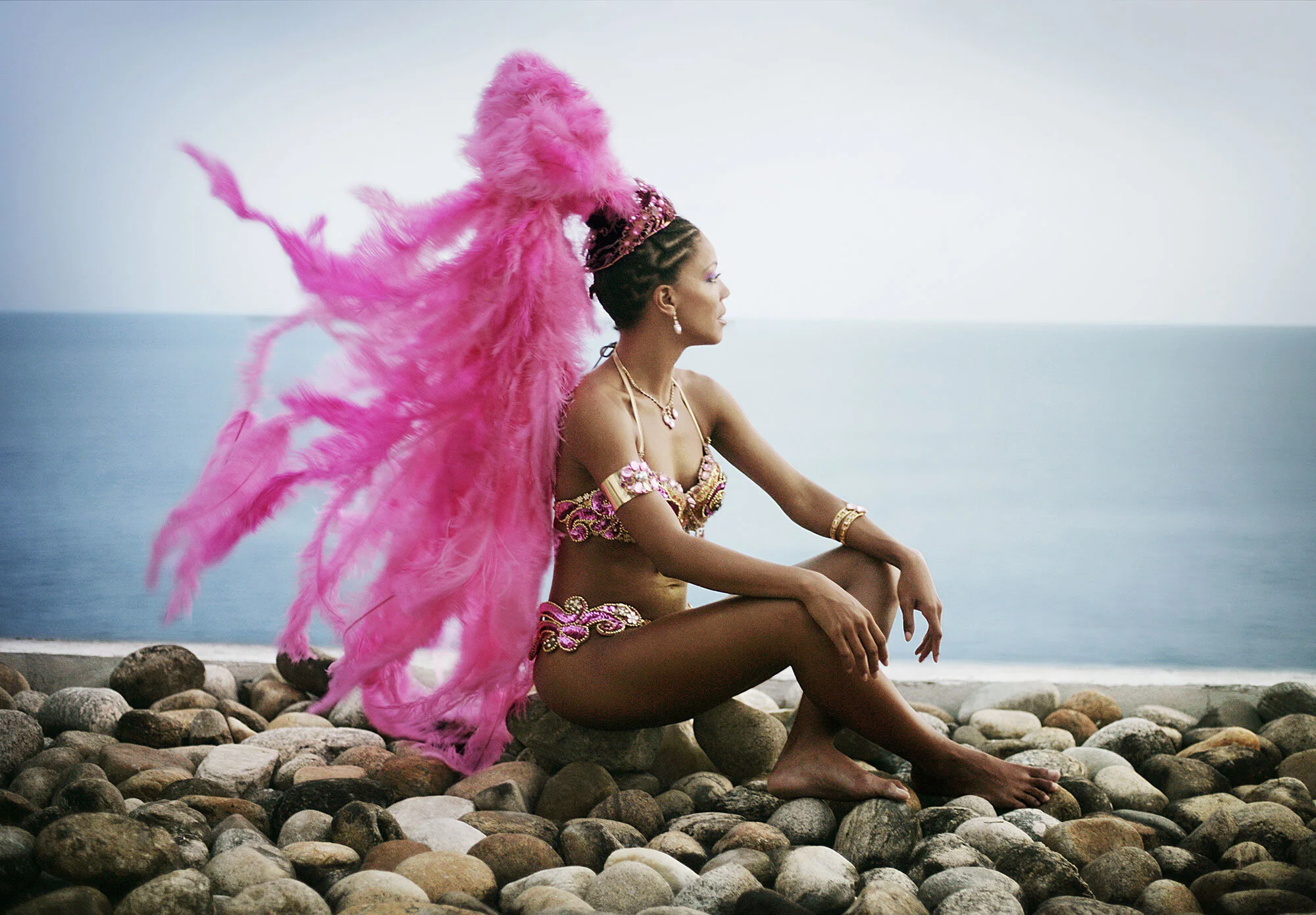 A Black Brazilian woman dressed in elaborate Carnival costume with pink feathers, jewelry, and a tiara, sitting on rocks by the sea with blue sky in Ipanema beach, facing in profile in Rio de Janeiro, Brazil.