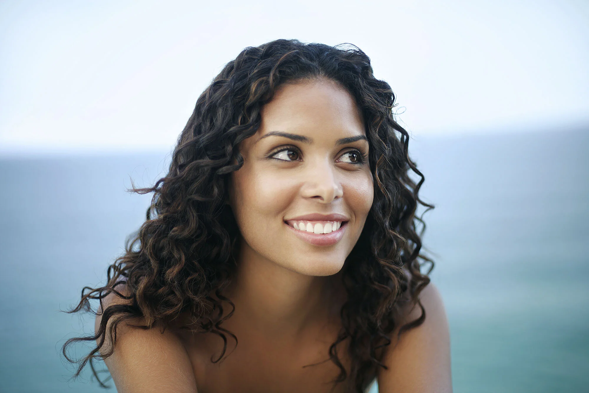 Close-up of a smiling Black Brazilian woman with curly brown hair looking to the side with an outdoor ocean backdrop of the Atlantic ocean at Ipanema beach in Rio de Janeiro, Brazil.