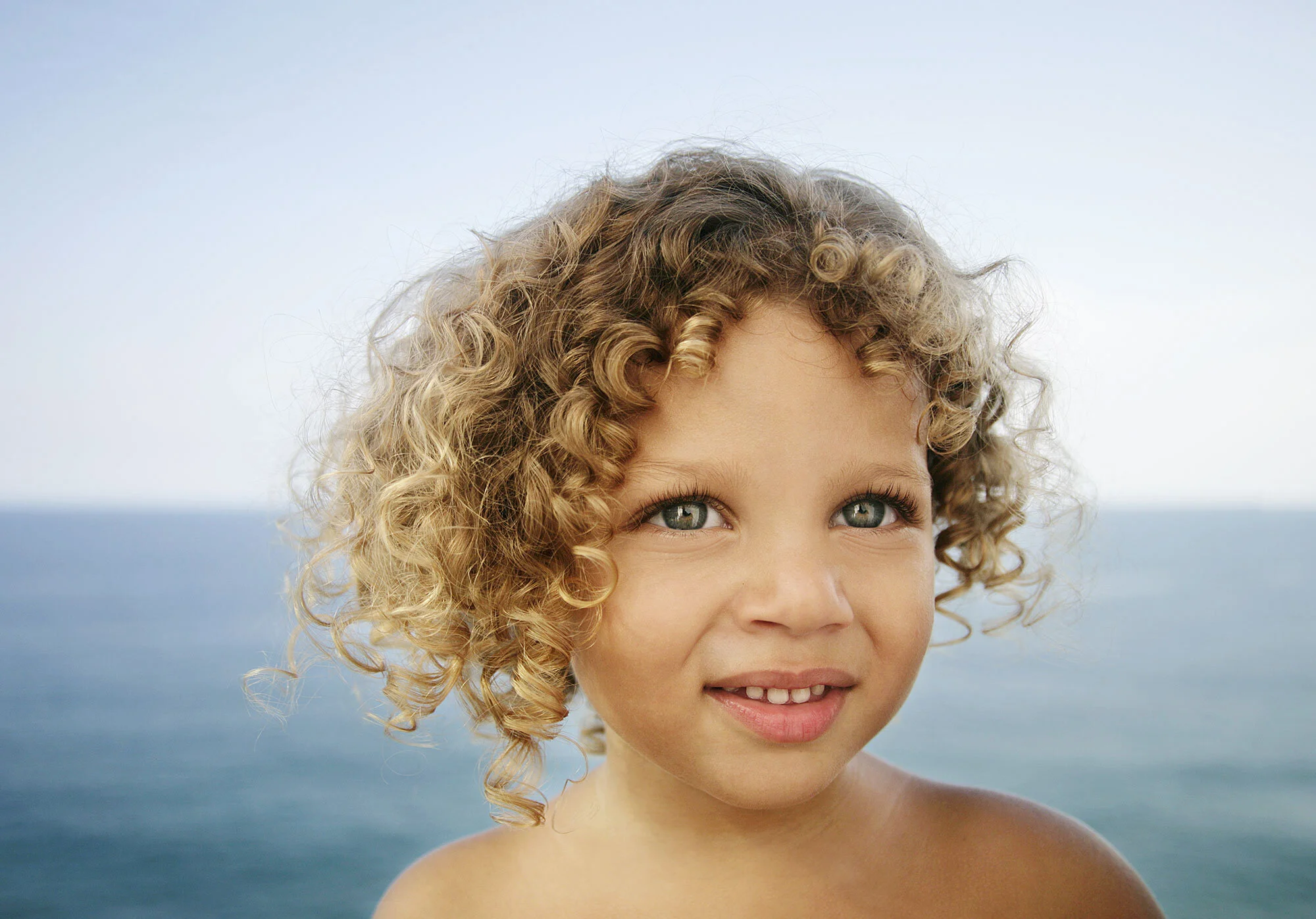 Close-up of a young girl with curly blonde hair and blue eyes outdoors with ocean in background at Ipanema beach in Rio de Janeiro, Brazil.