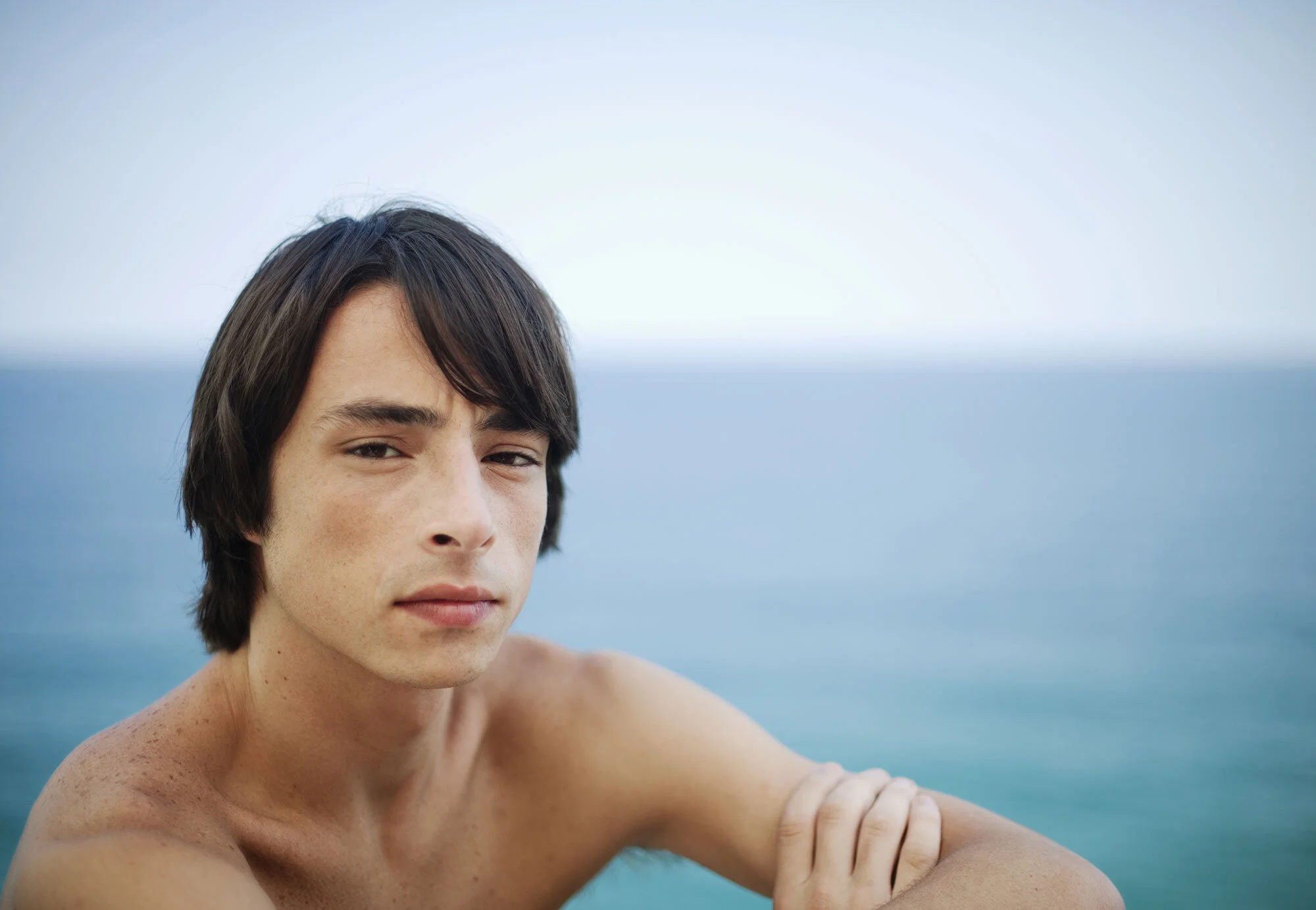 A young white Brazilian man with dark, straight hair and light freckles gazes at the camera against a backdrop of blue ocean and sky against the Atlantic ocean at Ipanema beach in in Rio de Janeiro, Brazil.