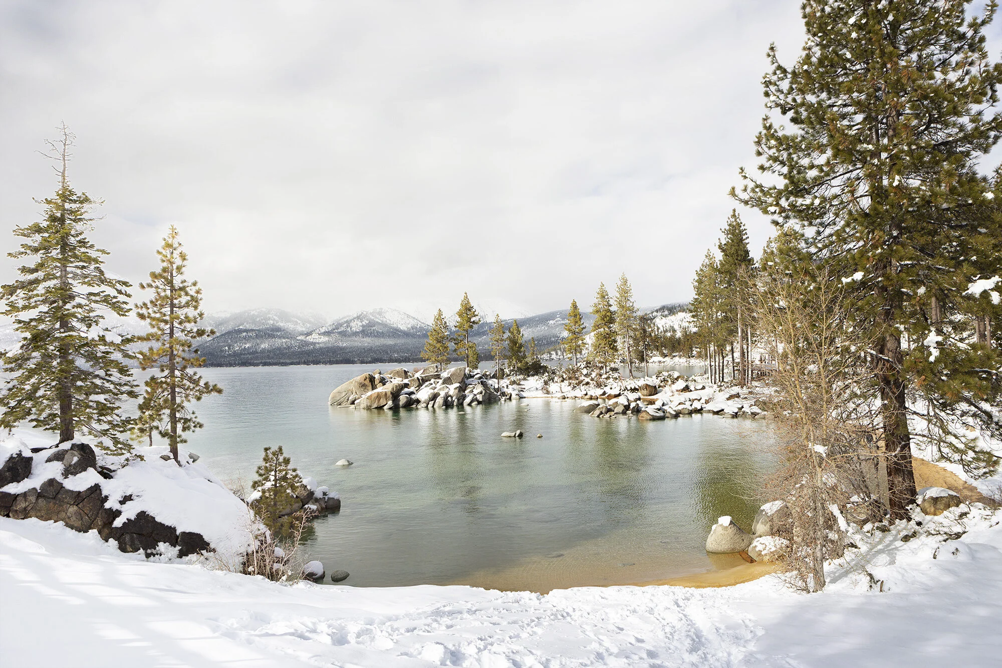 Snow-covered shoreline with pine trees and a calm lake in a mountainous landscape at Sand Harbor in Lake Tahoe, CA.