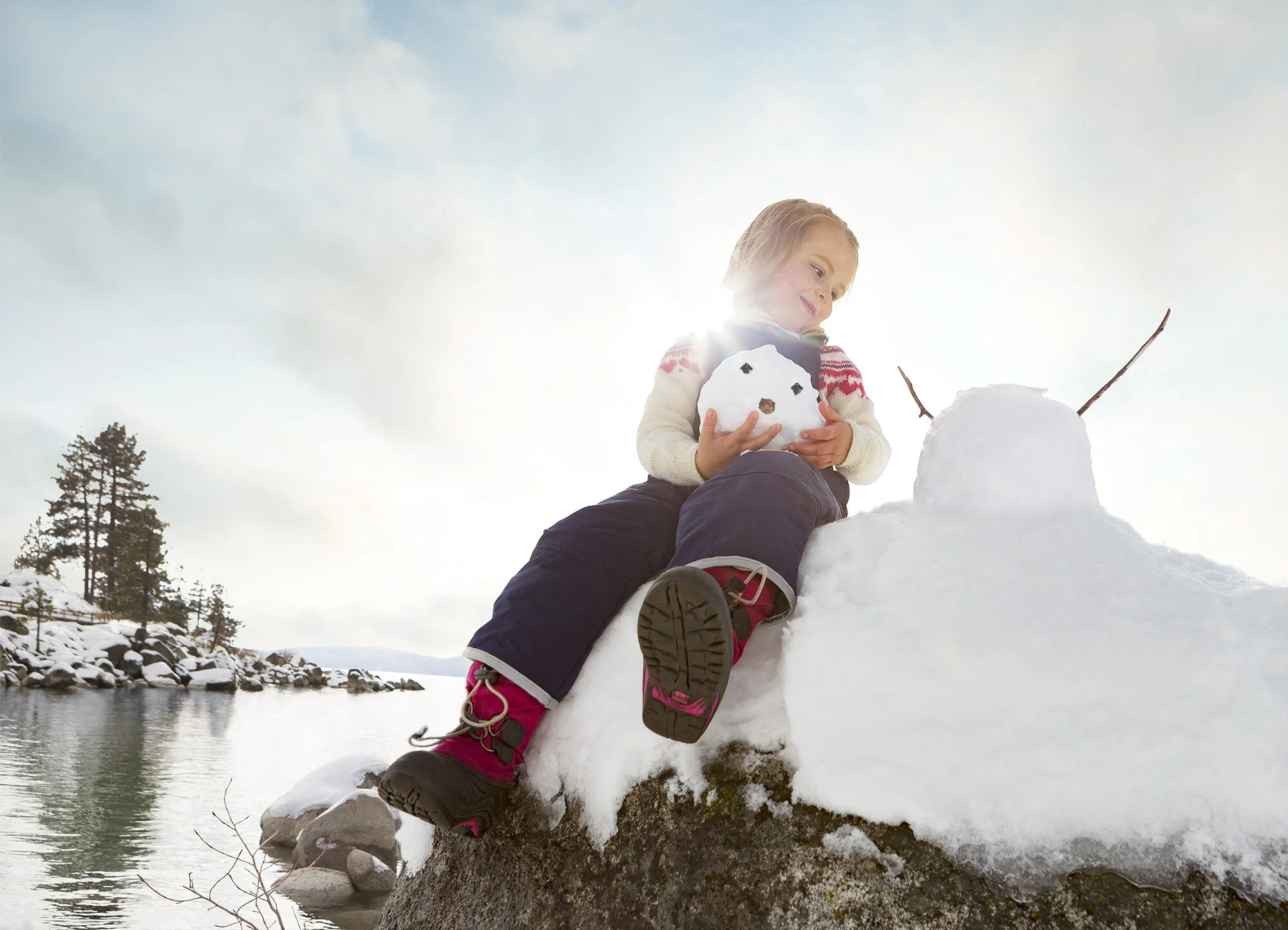 A young girl sitting on a large rock by the water, holding a small snowman with a surprised expression, a snow-covered landscape with trees and a cloudy sky in the background at Sand Harbor in Lake Tahoe, CA.