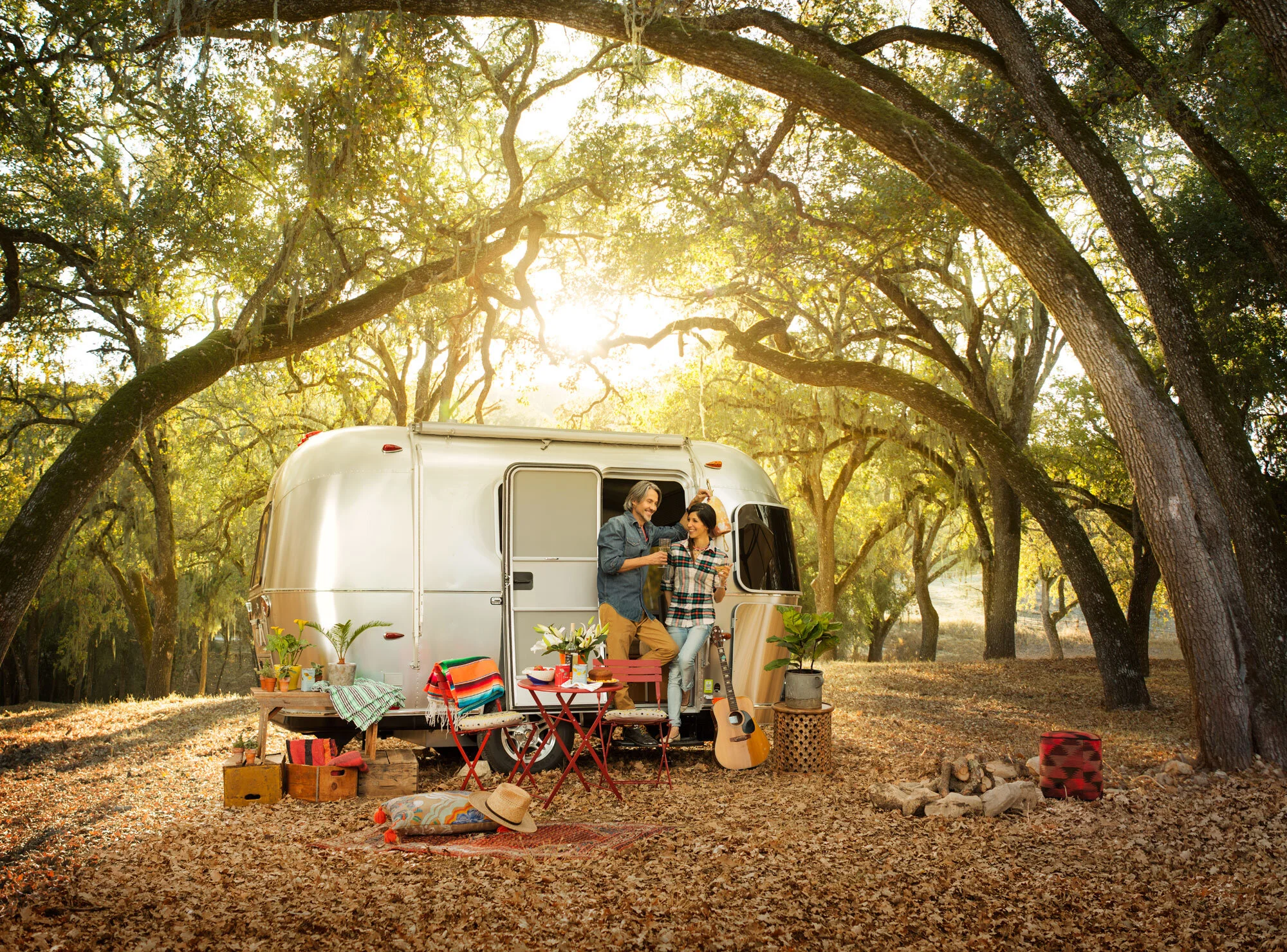 A Latino couple standing outside a camper van glamping in a wooded area during sunset under oak trees, enjoying drinks and smiling at each other. Table with flowers, a guitar, potted plants, a picnic blanket, and camper supplies in Paso Robles, CA.